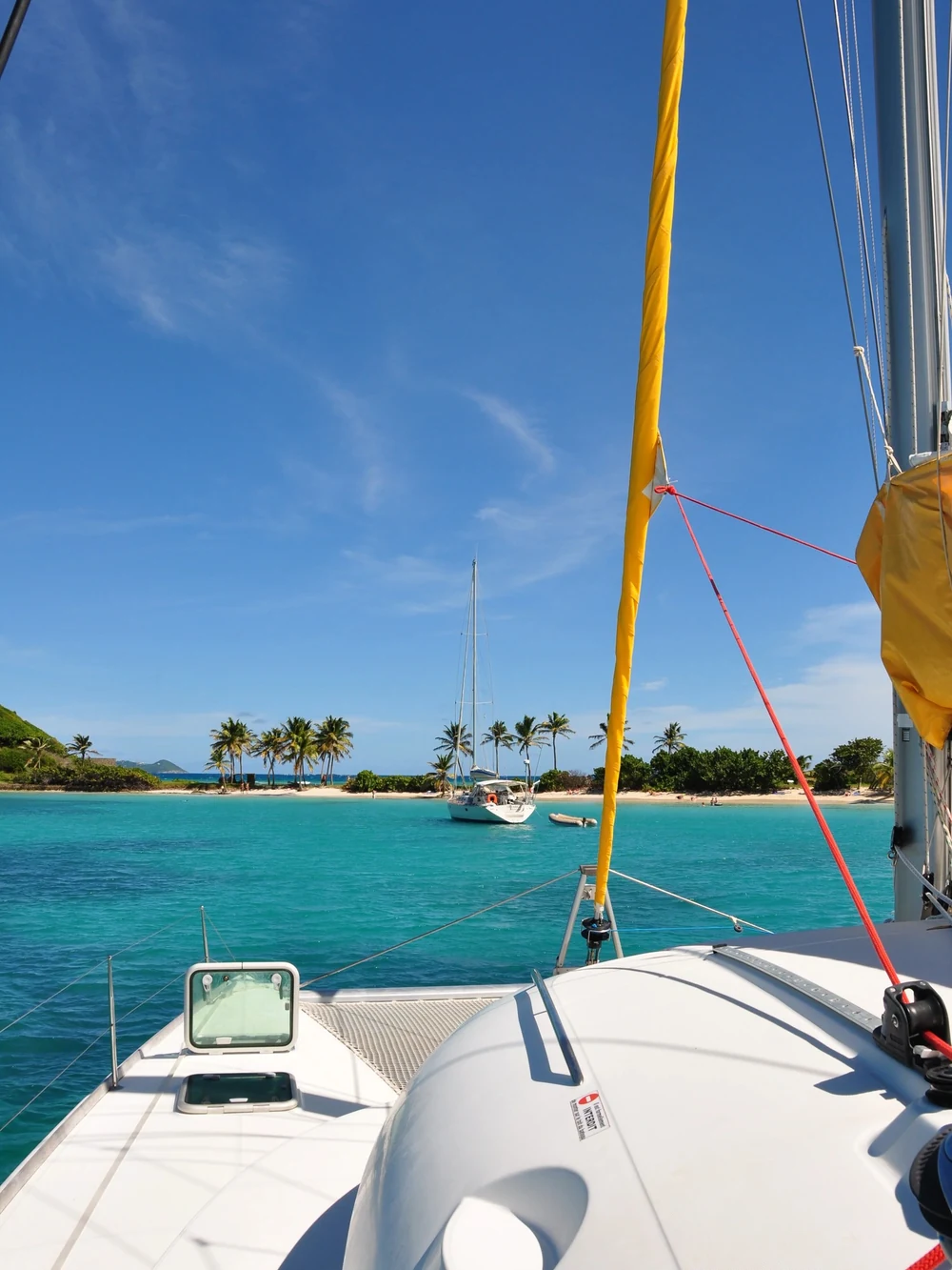 The bow of a private catamaran glides toward a palm-fringed island with another sailboat anchored offshore.