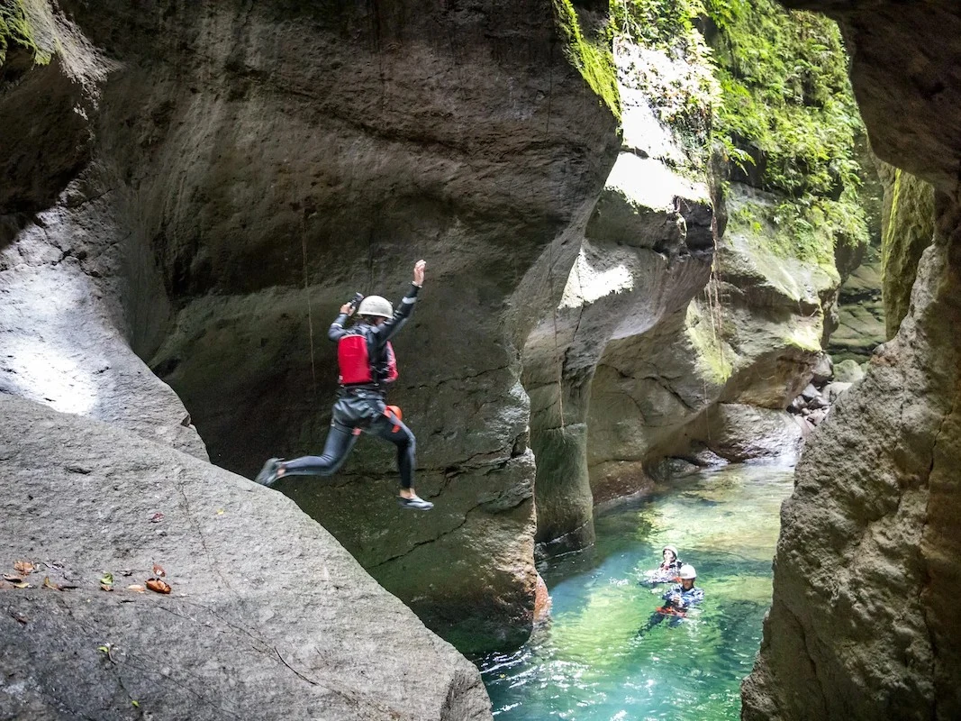 A canyoneer in a helmet and red life vest leaps off a rock ledge into a turquoise plunge pool deep in a Dominica canyon, while two guides tread water below.