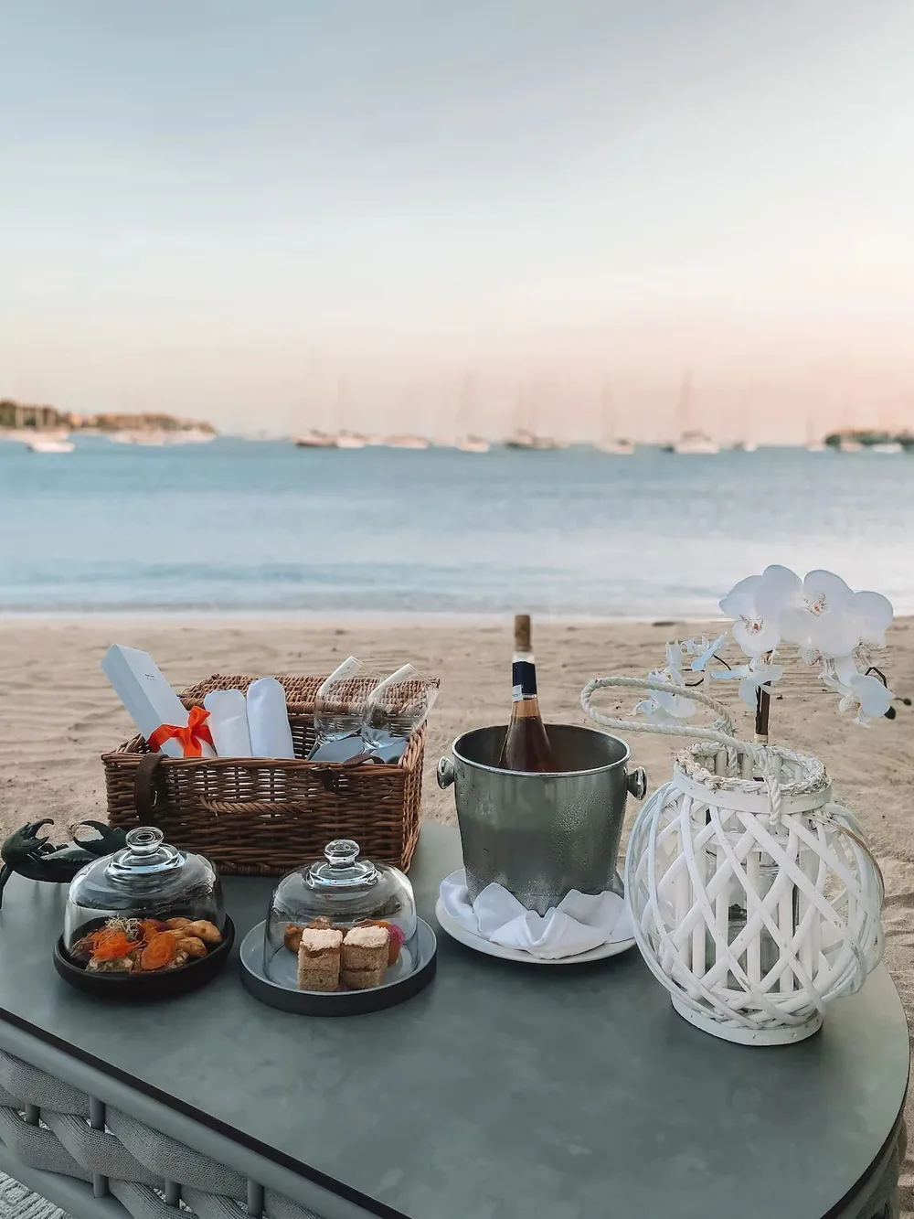 Beach picnic with food, drinks, and a lantern by the water.