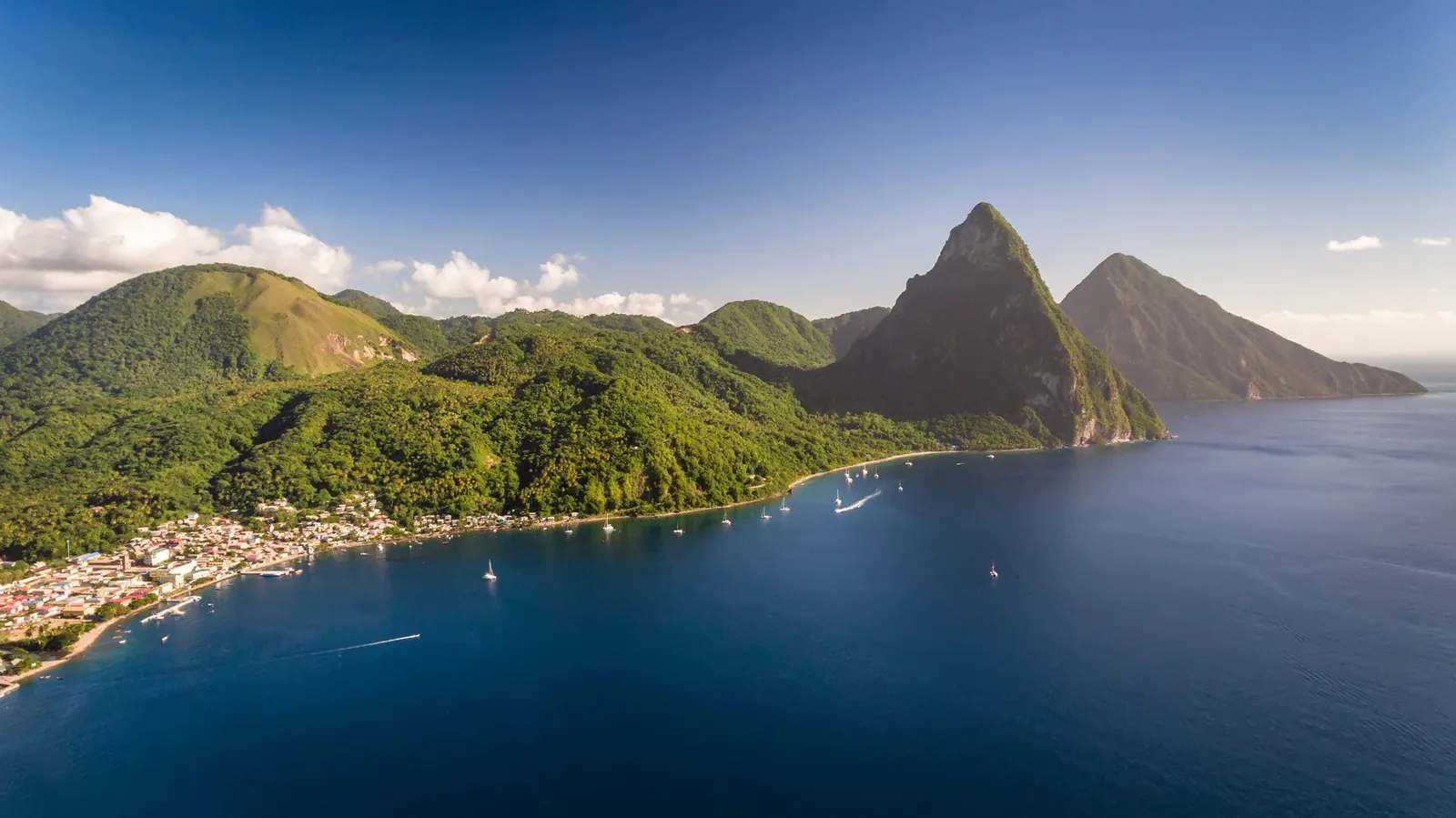 Aerial view of Soufrière Bay and the Pitons rising from the sea, with sailboats anchored along the forested coast.