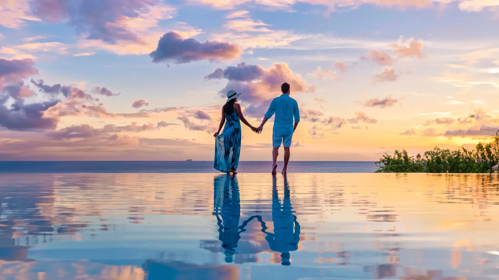 A couple walks hand in hand along the edge of an infinity pool at sunset, the sky reflected in the water.