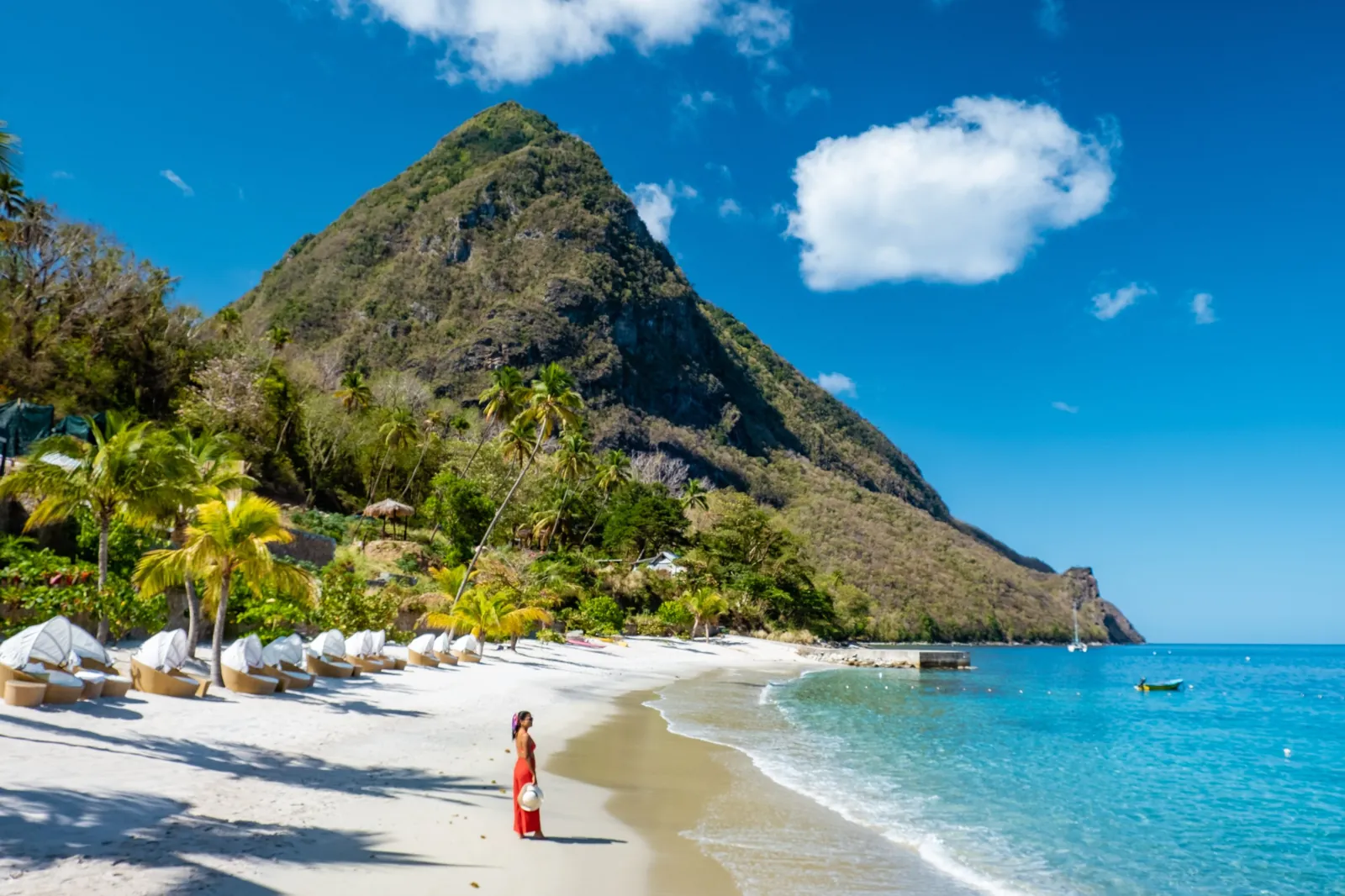 A woman in a red dress stands on a white-sand beach below Petit Piton, with turquoise water lapping the shore.