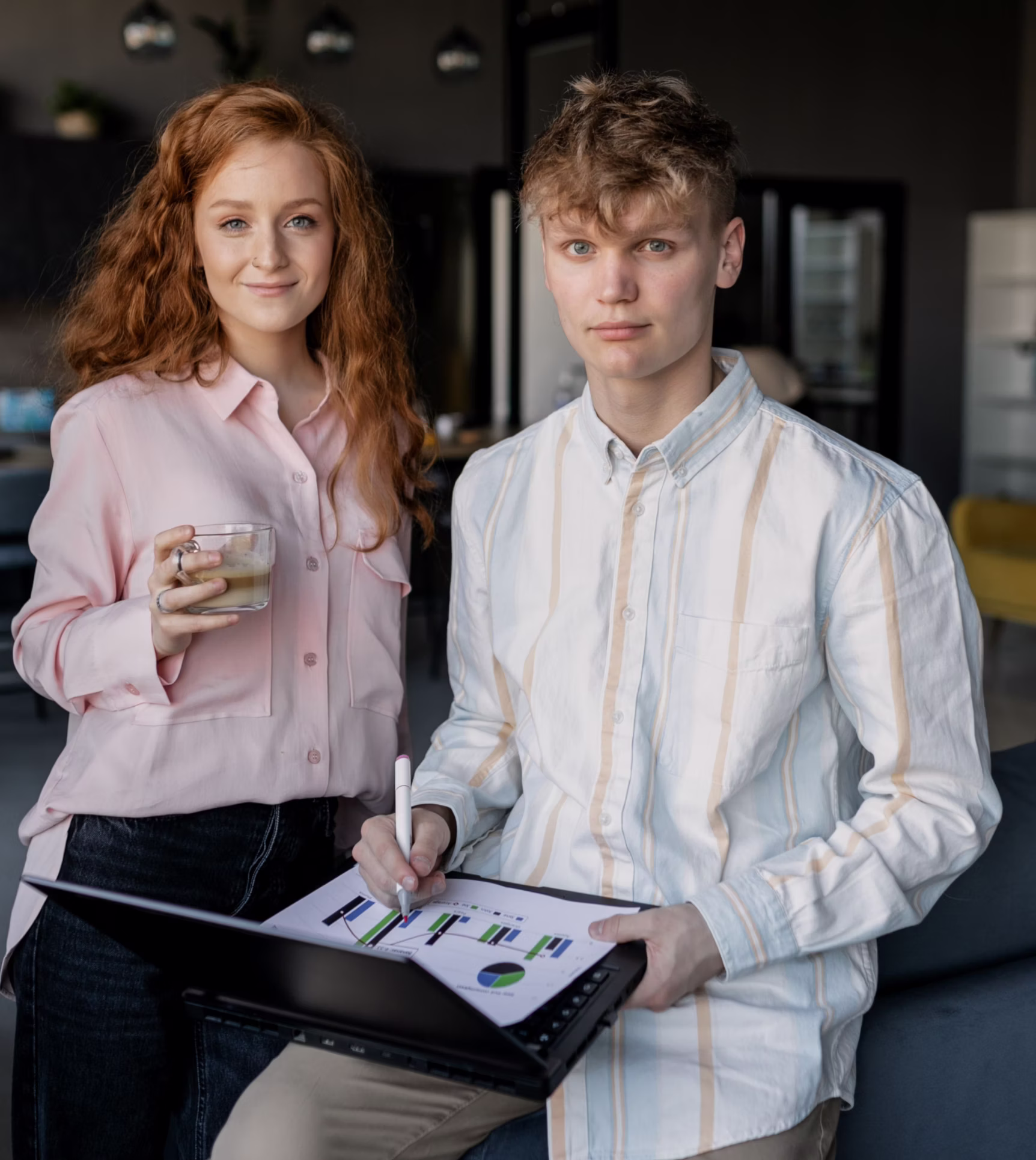 A young woman with long red hair holding a glass with a beverage stands next to a young man with short curly hair wearing a white striped shirt, who is seated and writing on a laptop displaying charts and graphs in a modern indoor setting.