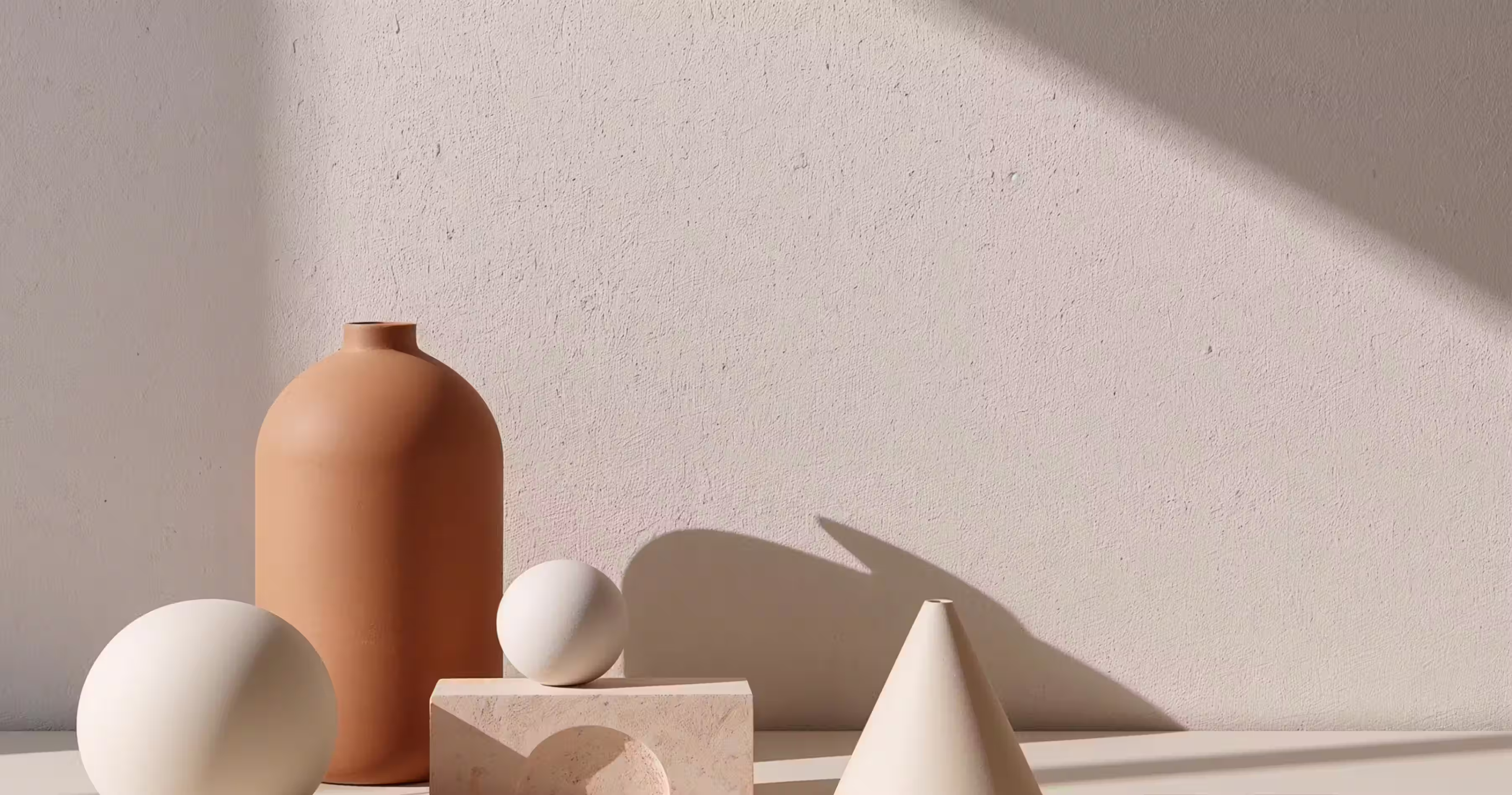 Minimalist arrangement of beige and white geometric objects including a tall rounded vase, two spheres, a cone, and a rectangular block cast soft shadows on a light textured wall.