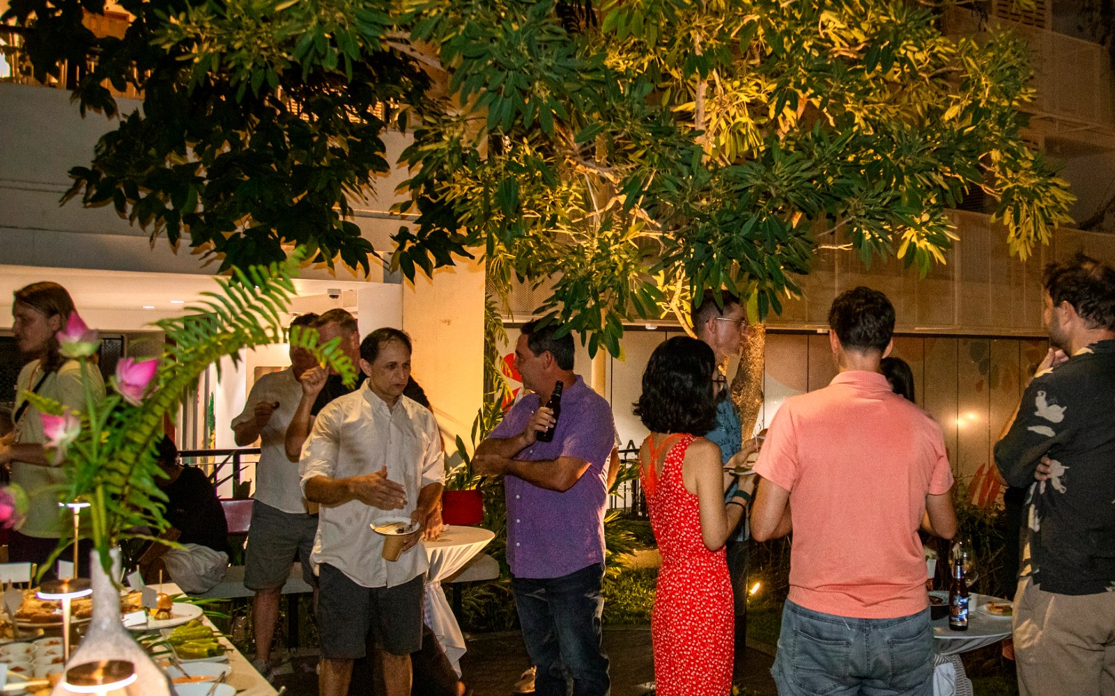 A private evening event at Commune, guests standing with drinks around a long table under the trees