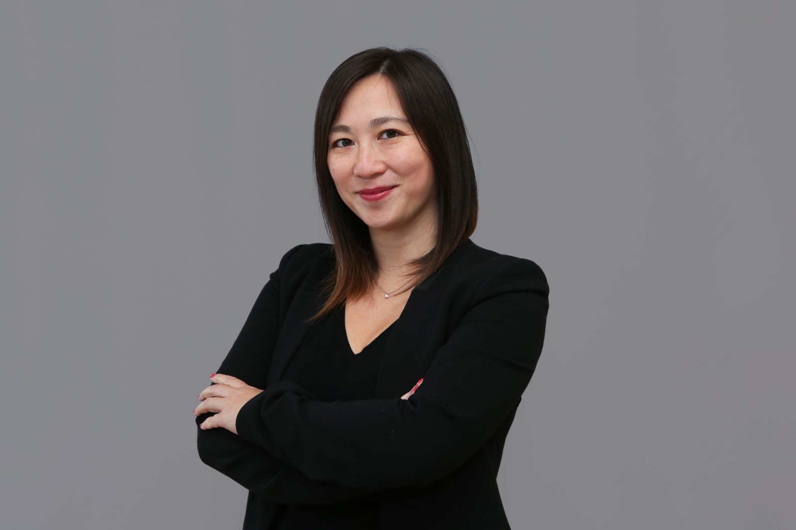 Confident woman with straight dark hair wearing a black blazer and necklace, smiling with arms crossed against a gray background.