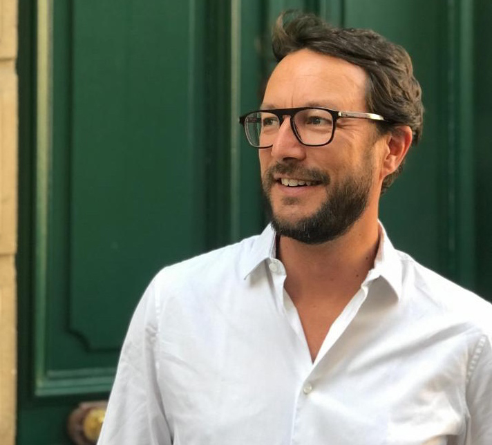 Man with dark hair, beard, and glasses wearing a white shirt standing in front of a dark green door, looking to his left and smiling.