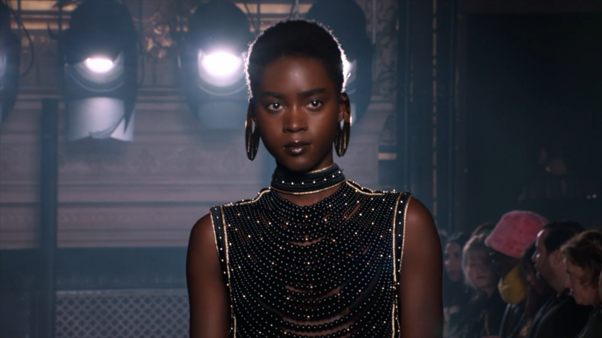 Model with short hair and large hoop earrings wearing a black dress adorned with small round beads, standing under bright stage lights with an audience in the background.