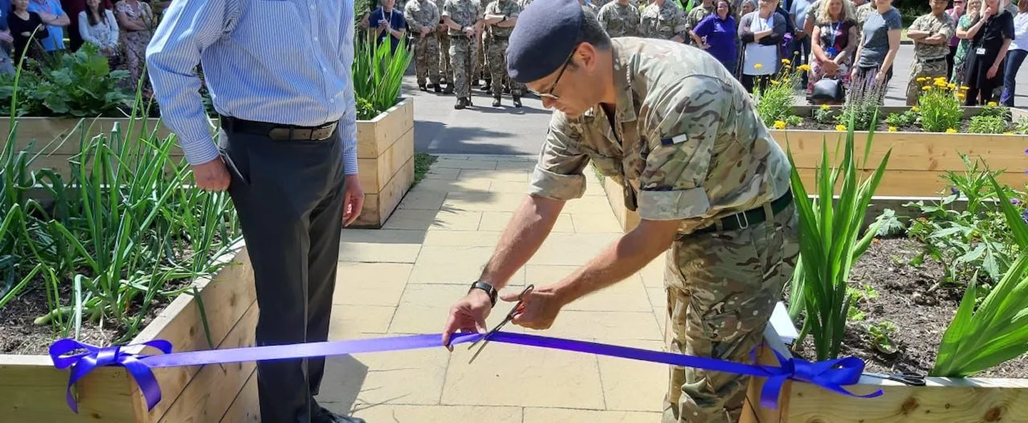Soldier in beret cutting a blue ribbon to open a garden with raised planters, watched by a crowd