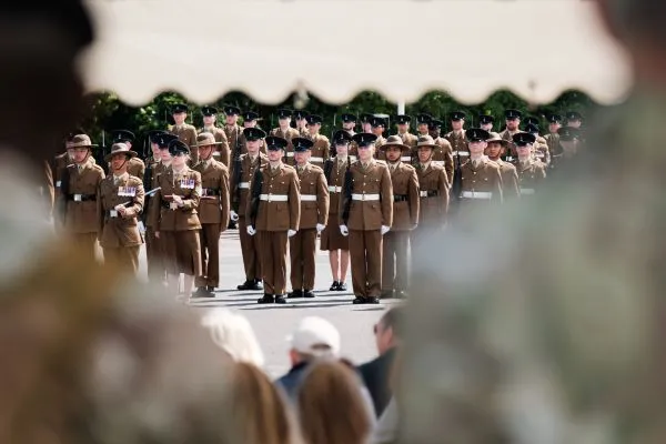 Soldiers in khaki dress uniform standing in ranks on a parade square, seen past blurred spectators
