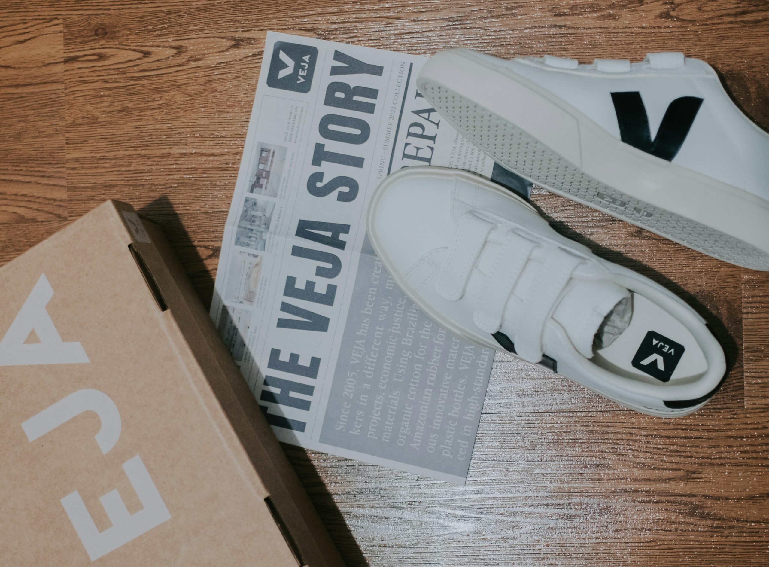 Zapatillas VEJA blancas, su caja y el folleto The VEJA Story sobre una superficie de madera.