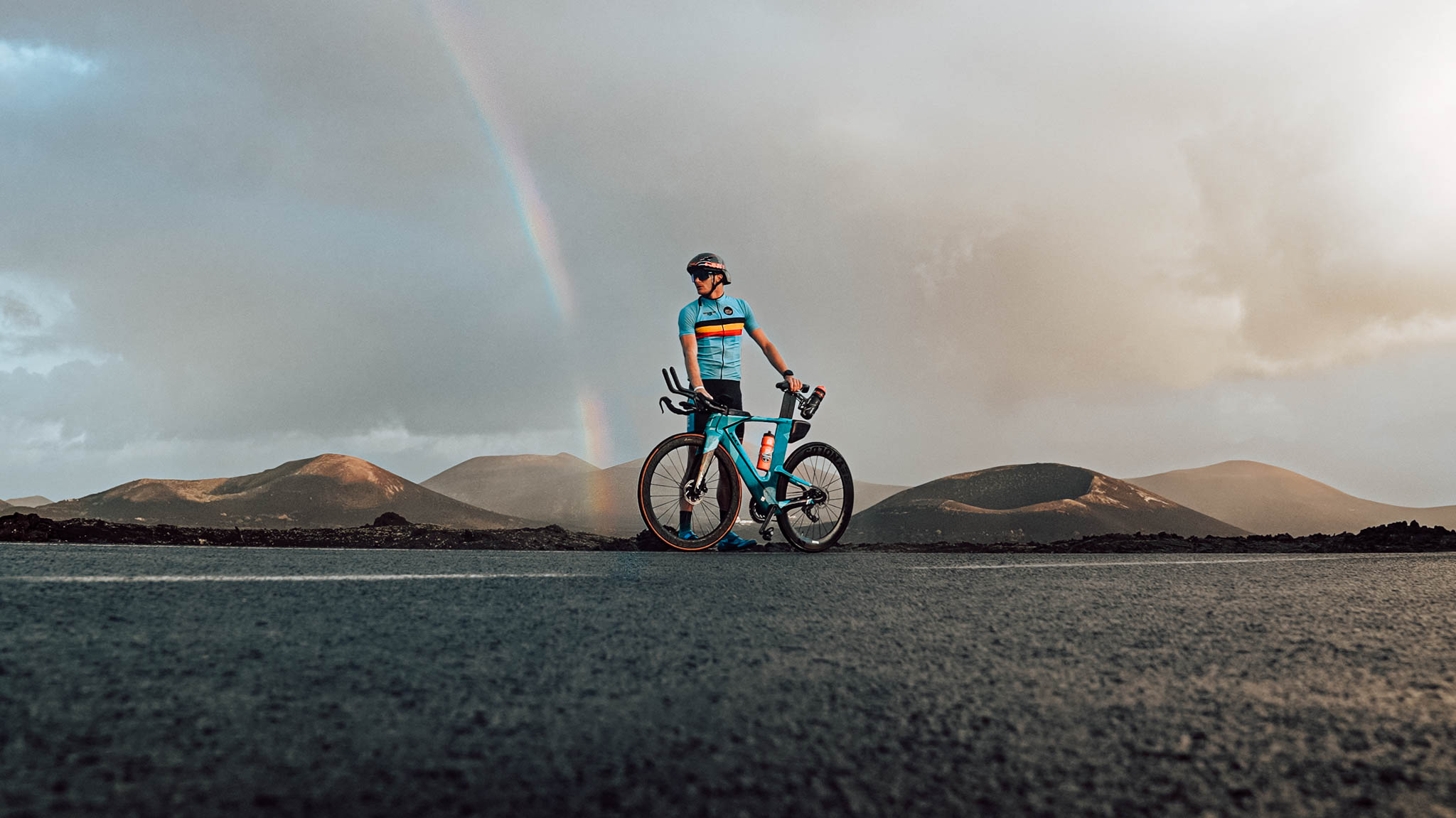Cyclist with a bike on a road in a volcanic landscape under a rainbow