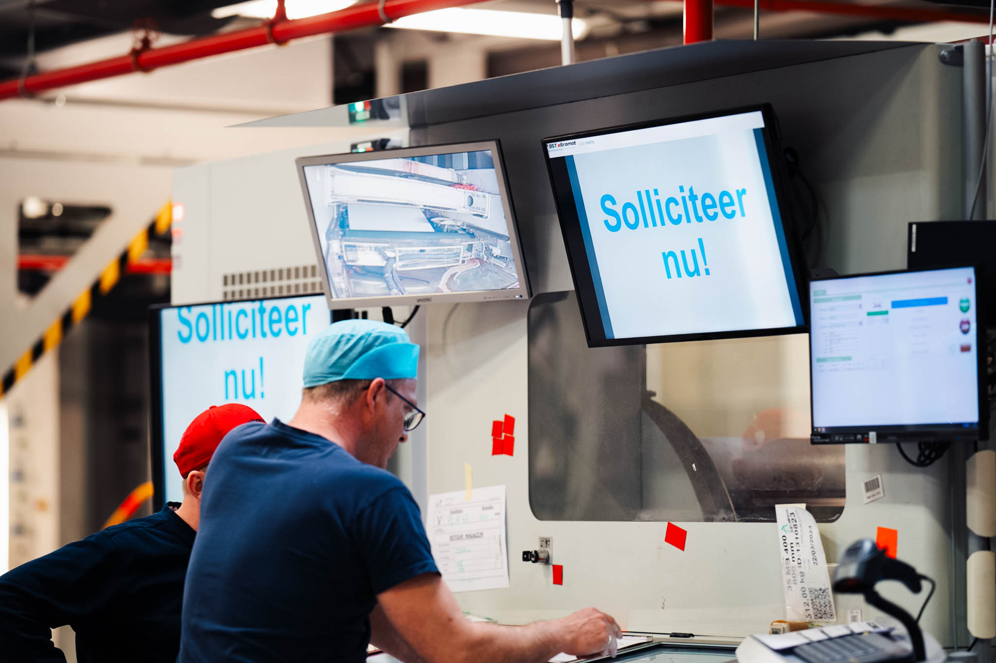 Factory workers at a machine with a 'Solliciteer nu' recruitment screen