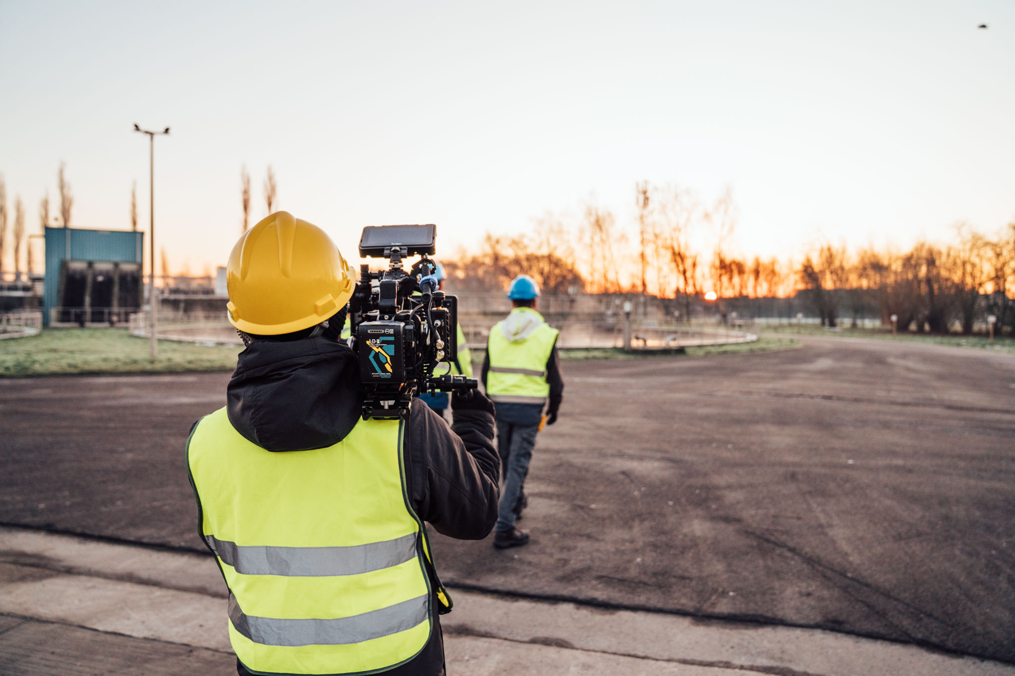 Camera operator in high-visibility gear filming on an industrial site at sunrise