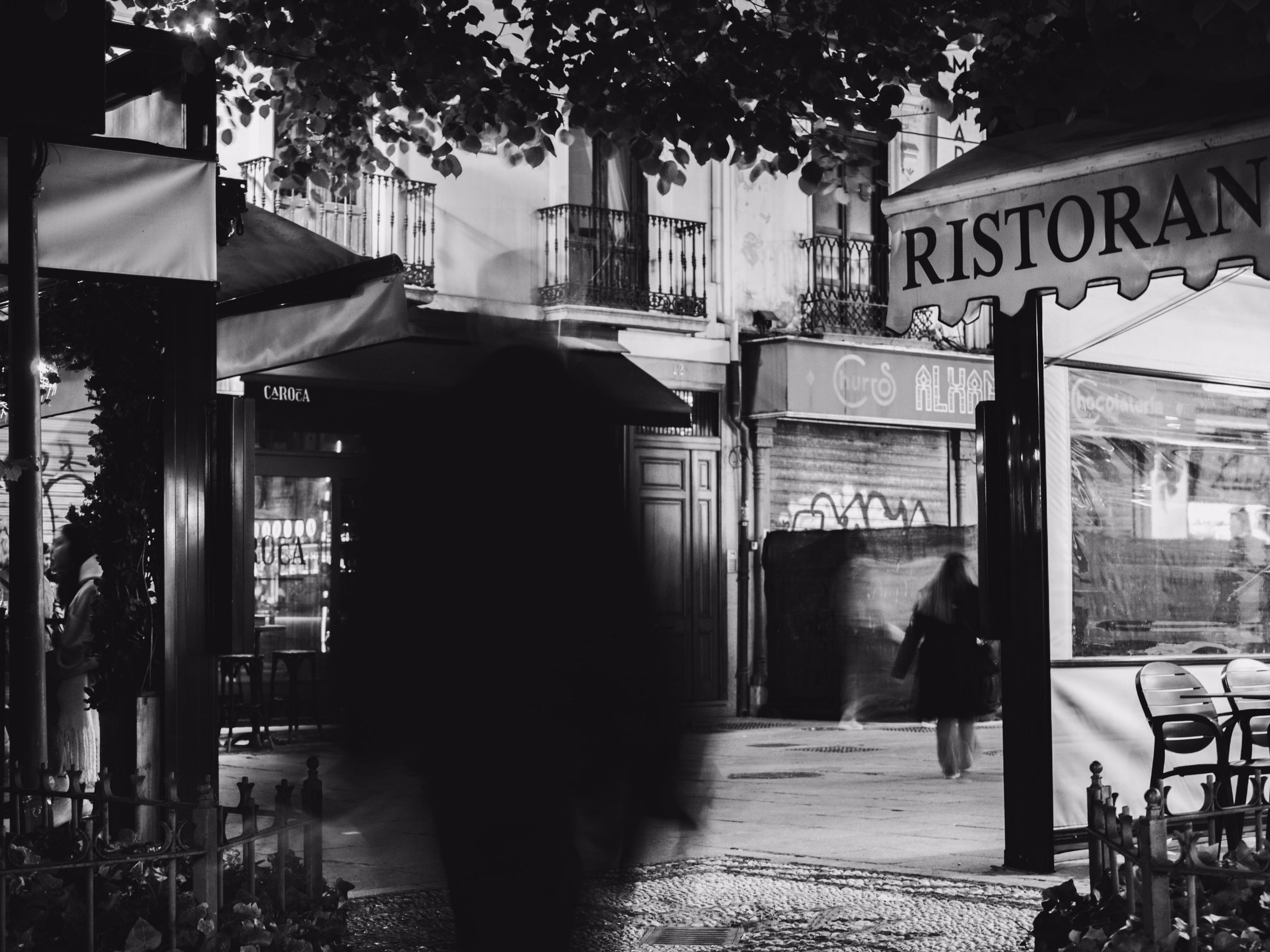 A blurred passerby walking past a ristorante sign on a city street, in black and white