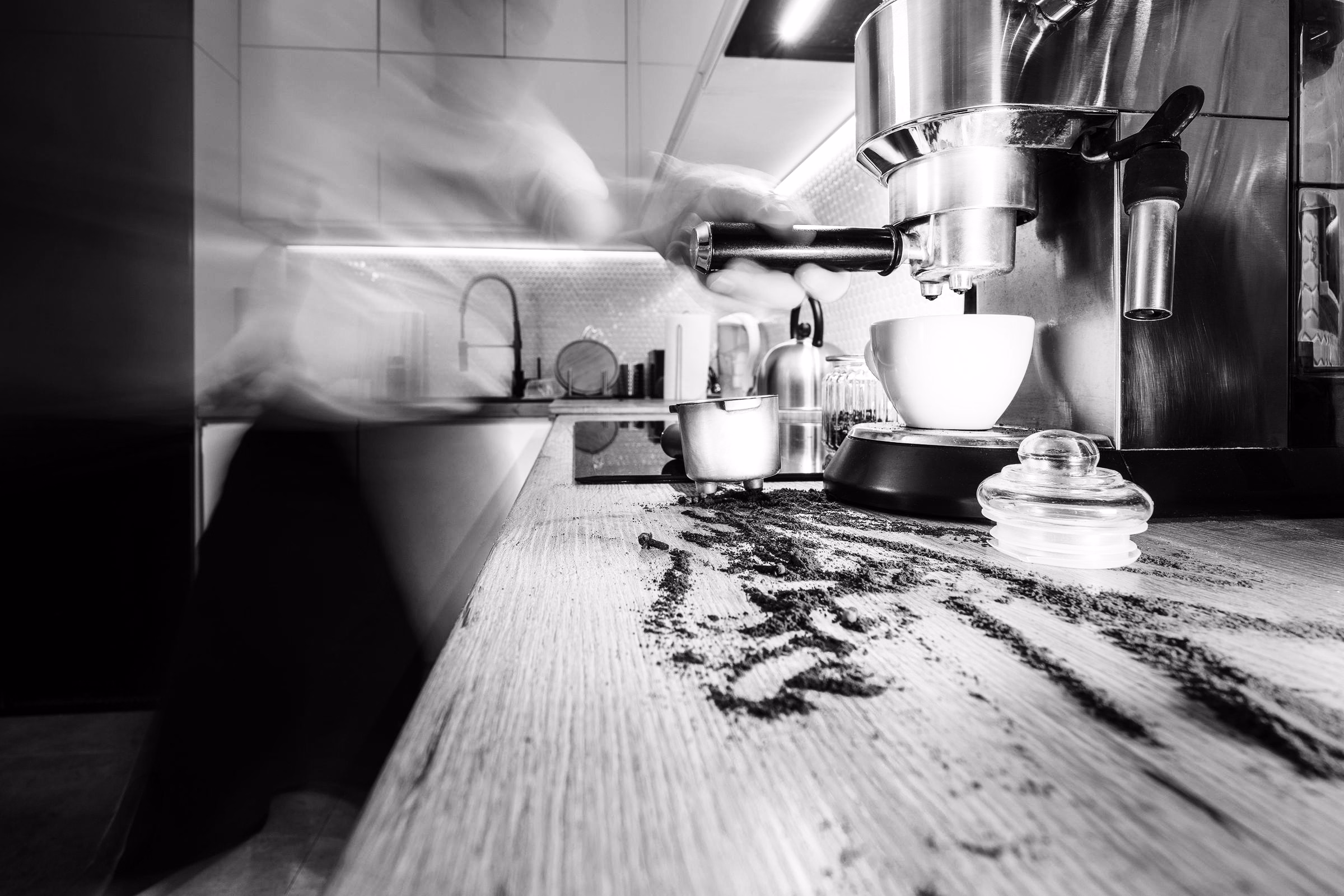 A blurred figure working behind an espresso machine at a cafe counter, in black and white