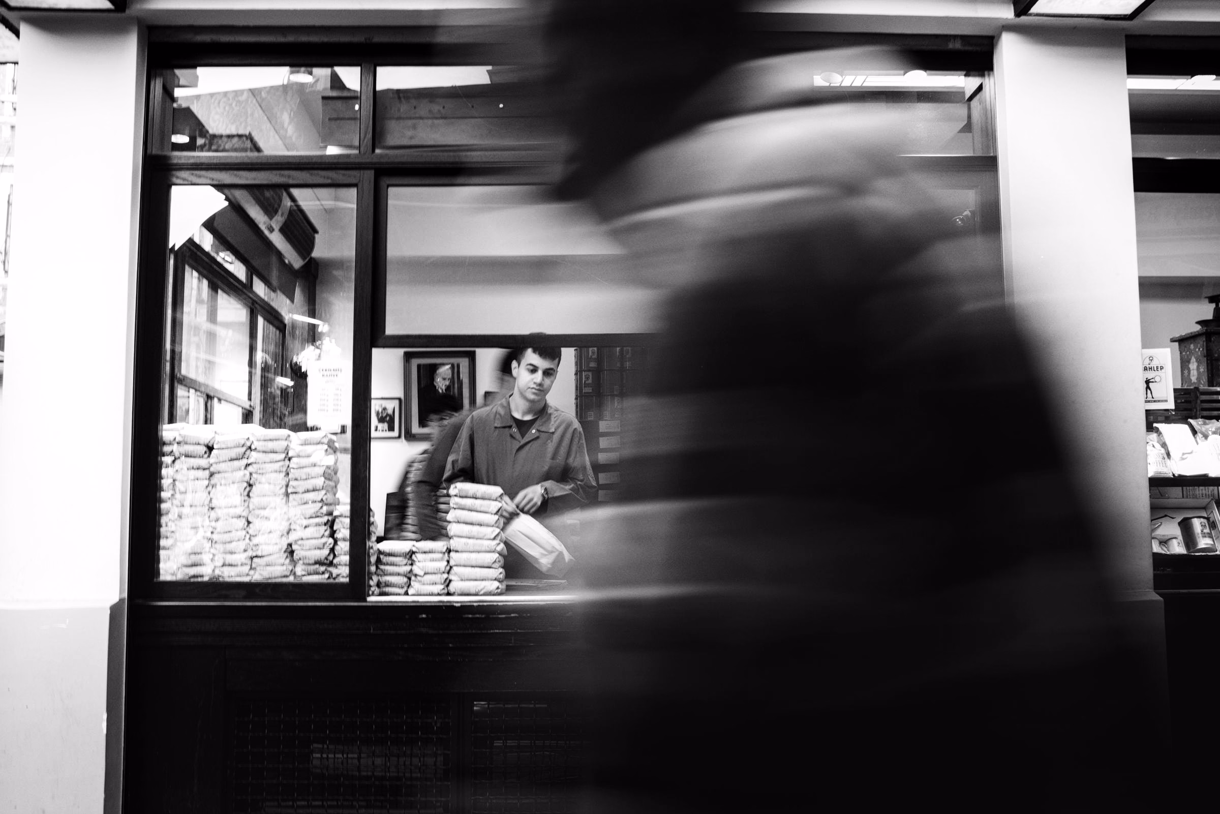 A blurred pedestrian passing a bakery window while a vendor works inside, in black and white