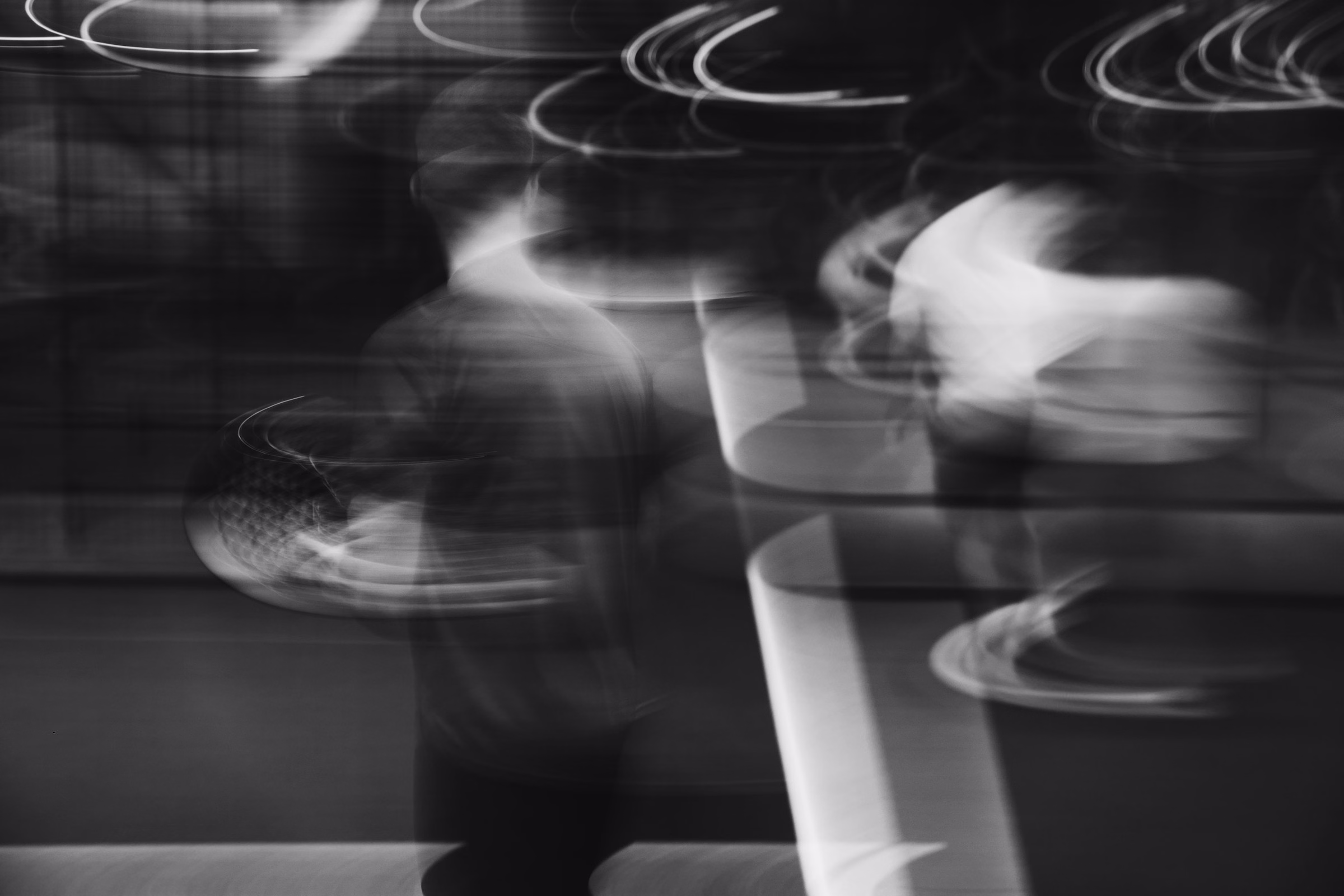 Blurred figures at a counter with arcs of light trailing above them, in black and white