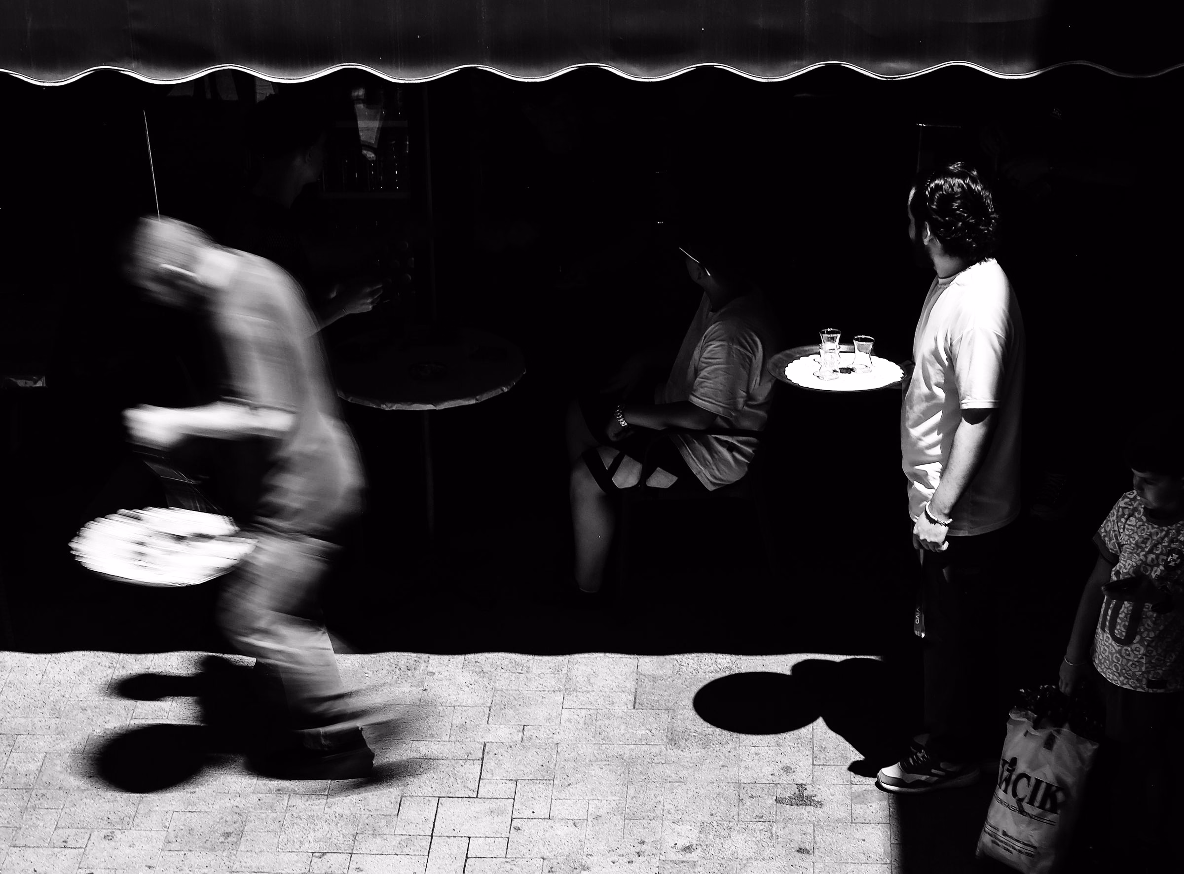A waiter hurrying across a terrace at night carrying plates, blurred in motion, in black and white