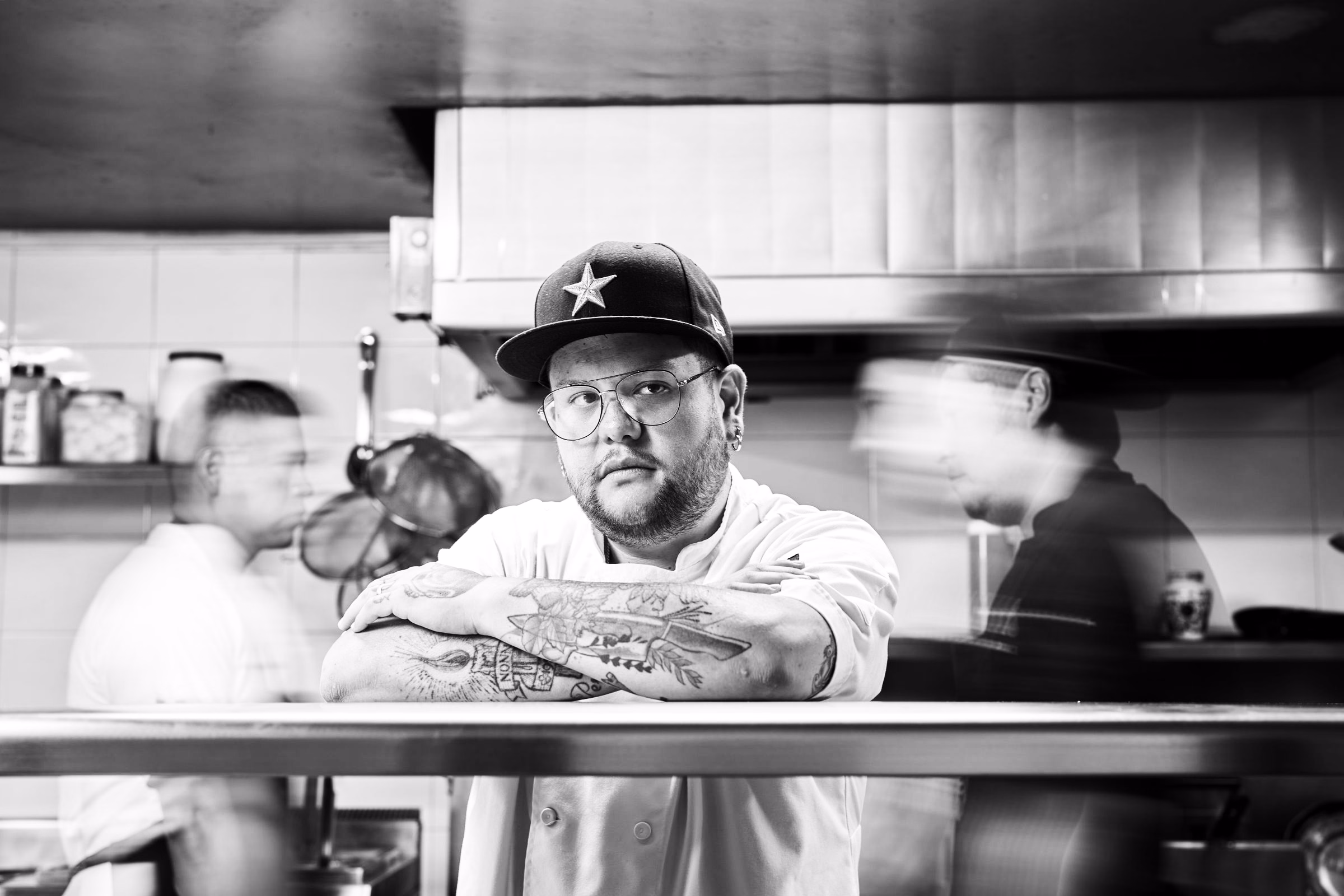 A chef standing still with arms crossed while the kitchen team blurs past, in black and white