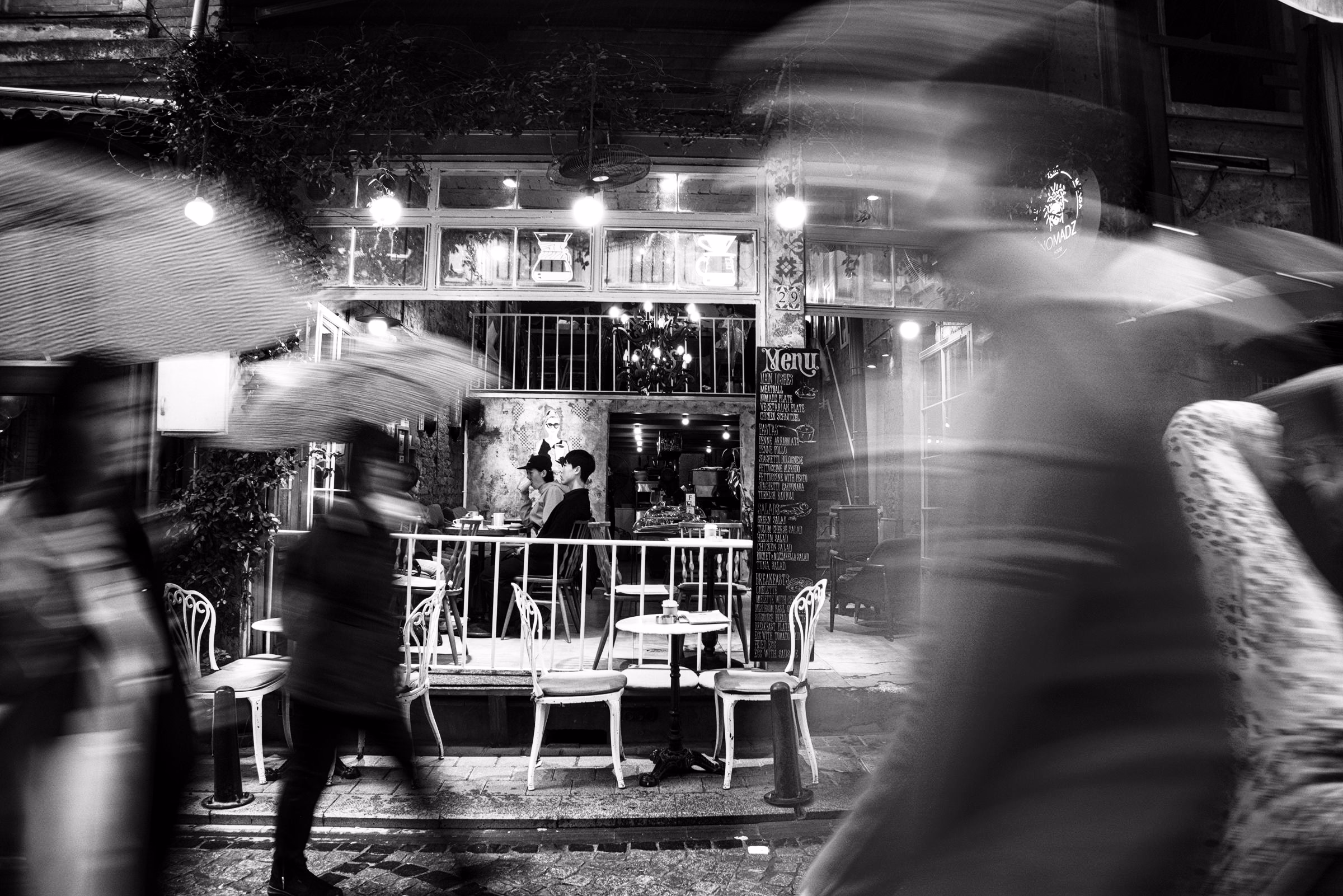 A lit cafe front at night with blurred people passing under umbrellas, in black and white