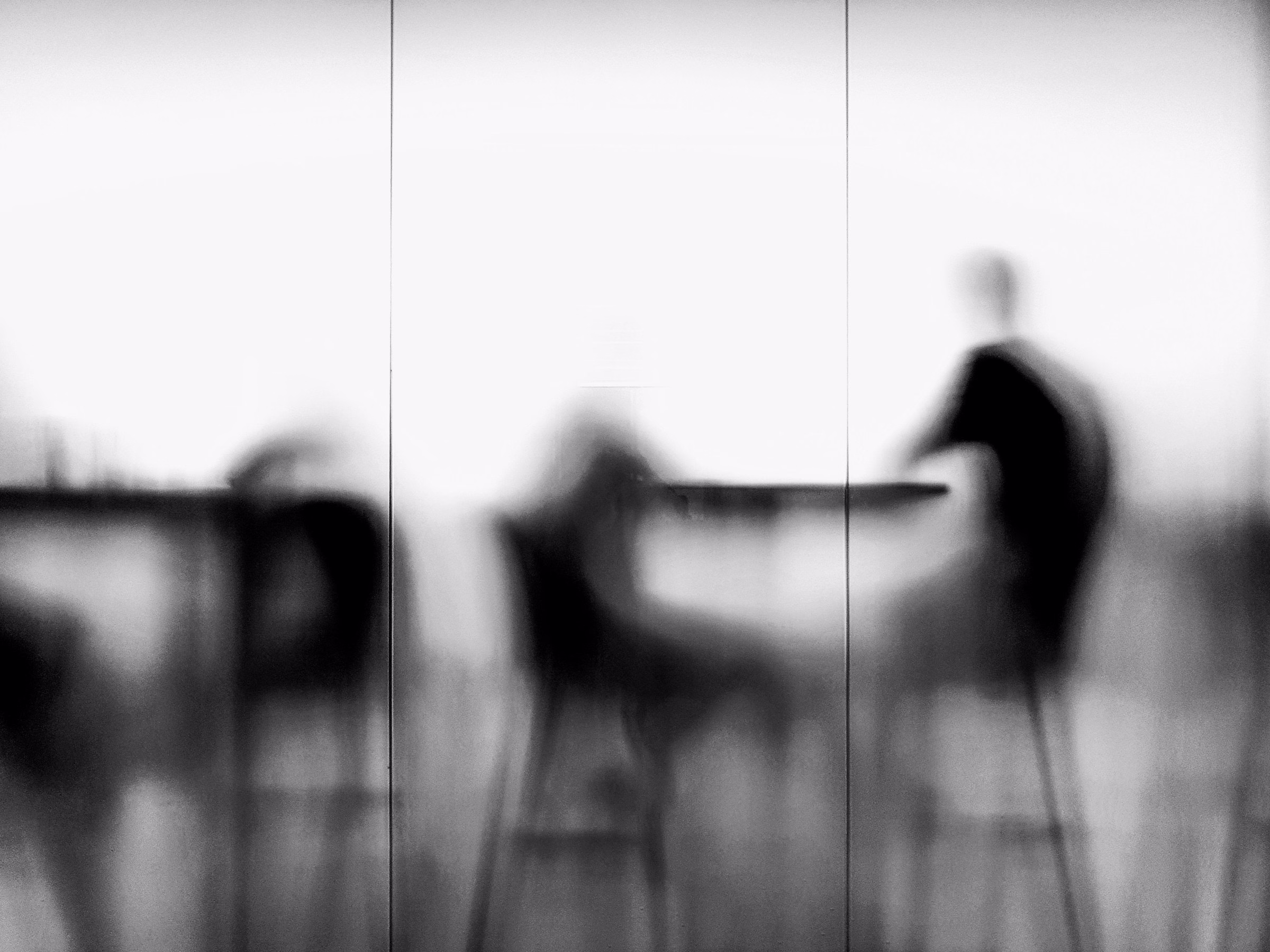 Blurred silhouettes of guests seated on stools at a cafe bar against a bright wall, in black and white