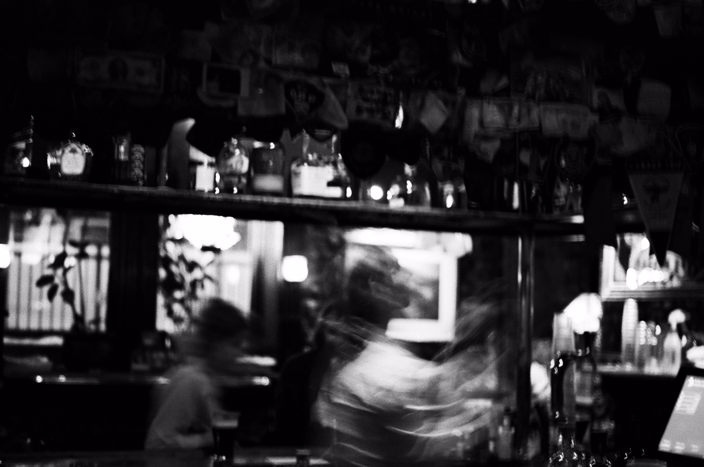 A blurred bartender moving behind a dimly lit bar counter, in black and white