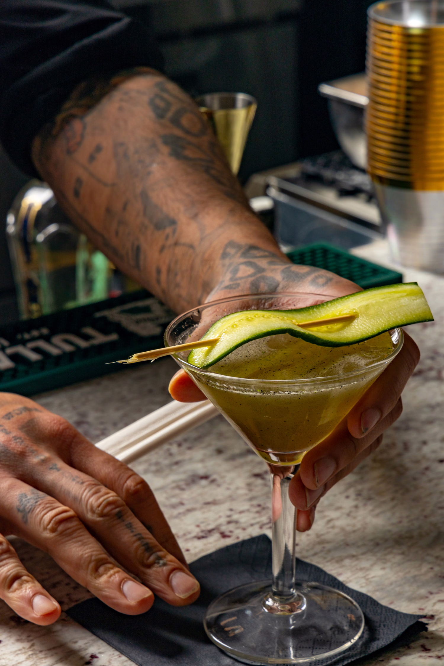 A green cocktail with a cucumber ribbon being set down on the bar at Auburn Angel