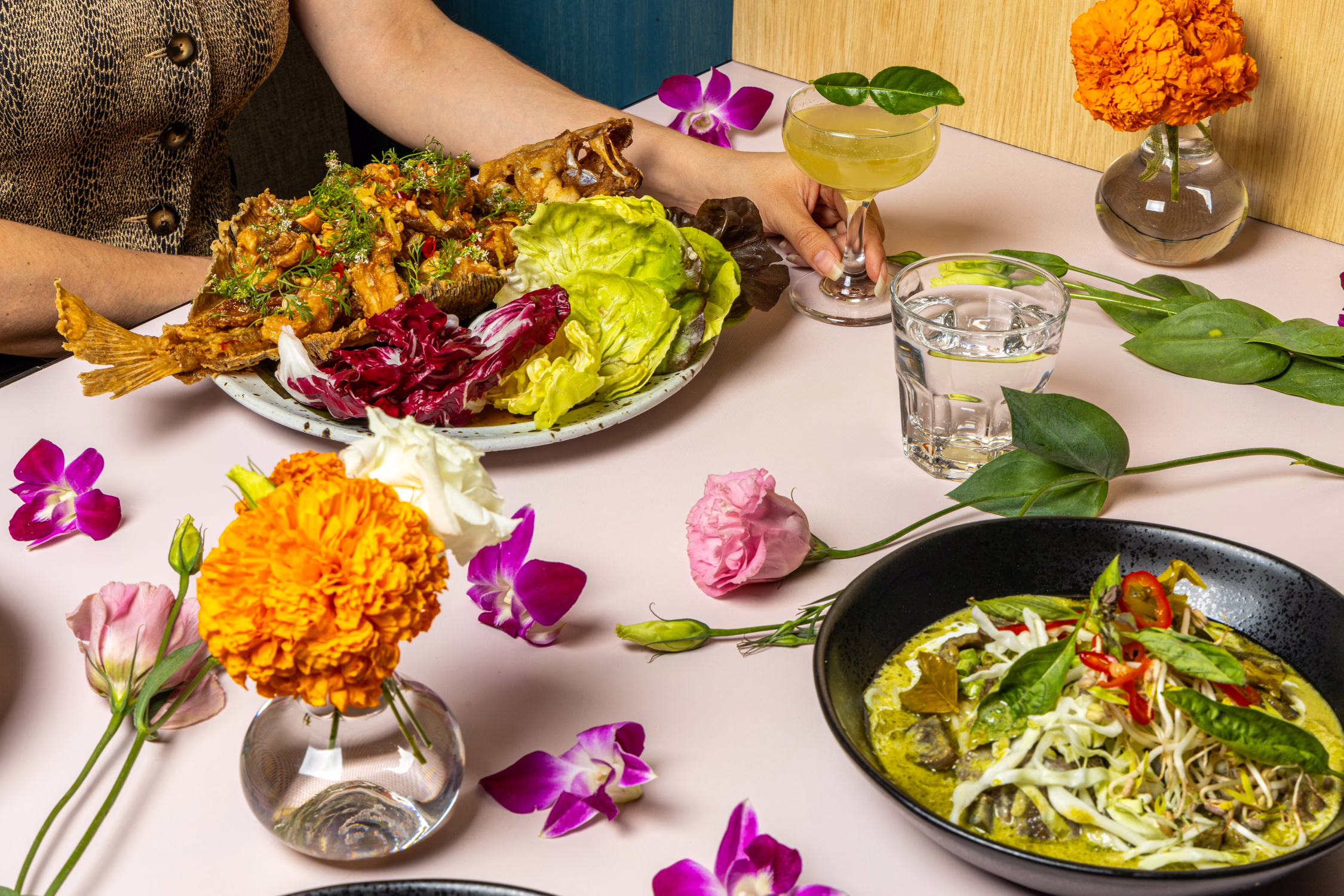 A table of whole fried fish, green curry noodles and a cocktail among flowers at Jo's Modern Thai