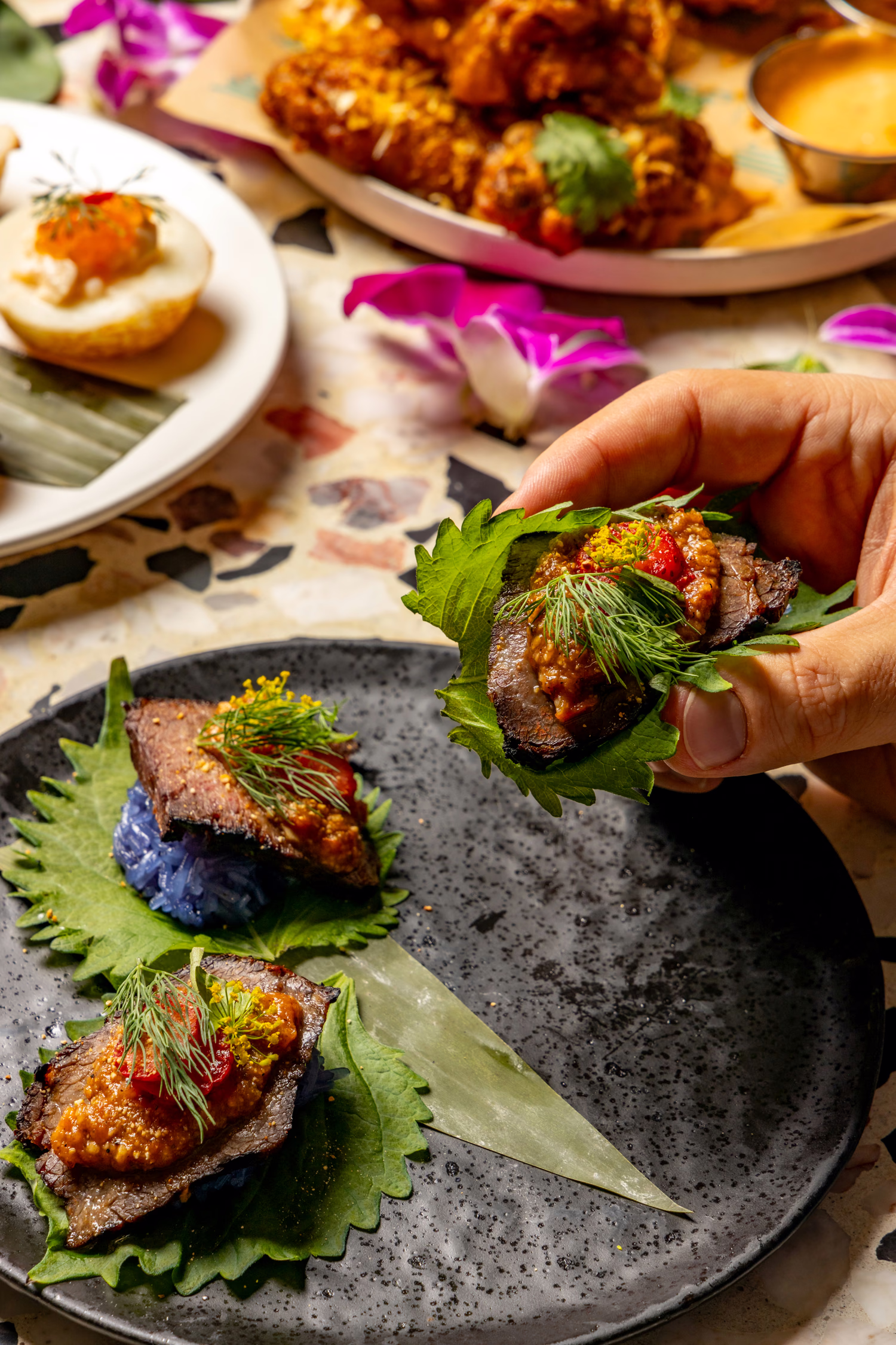 A hand lifting a leaf-wrapped bite from a dark plate at Jo's Modern Thai