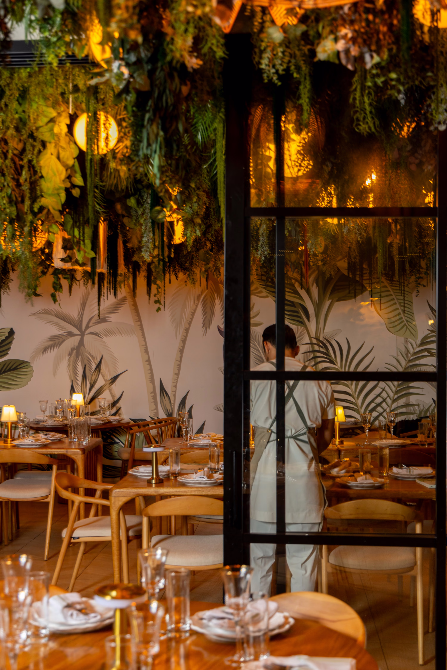 The dining room at Verdure seen through a glass door, with hanging greenery and candlelit tables