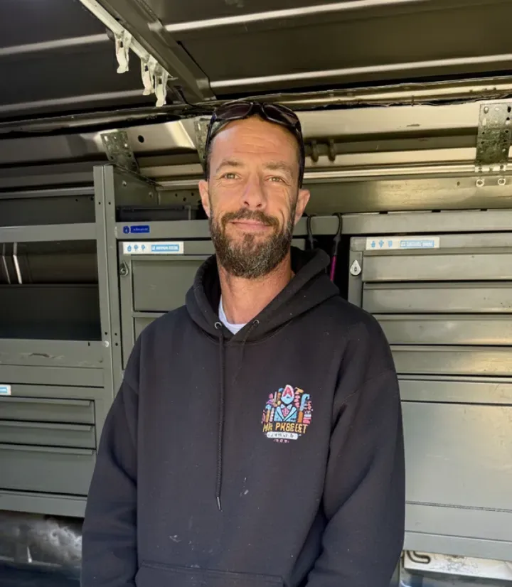 Man with short dark hair, beard, and sunglasses on his head wearing a black hoodie with a colorful logo standing inside a vehicle with metal storage cabinets.