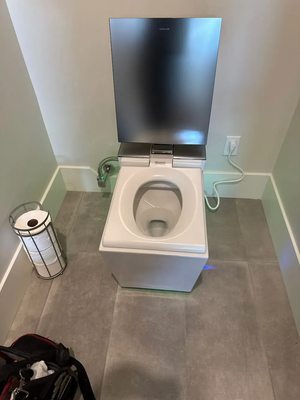 Modern white toilet with a stainless steel lid and lid cover in a tiled bathroom corner with a toilet paper holder containing multiple rolls on the left and an electrical outlet on the right wall.