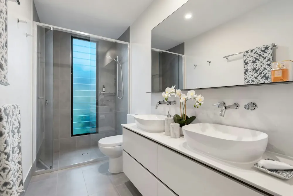 Modern bathroom with white double vessel sinks on a white vanity, large mirror above, wall-mounted faucets, a glass-enclosed shower with a frosted vertical window, toilet, and patterned gray and white towels on the wall.