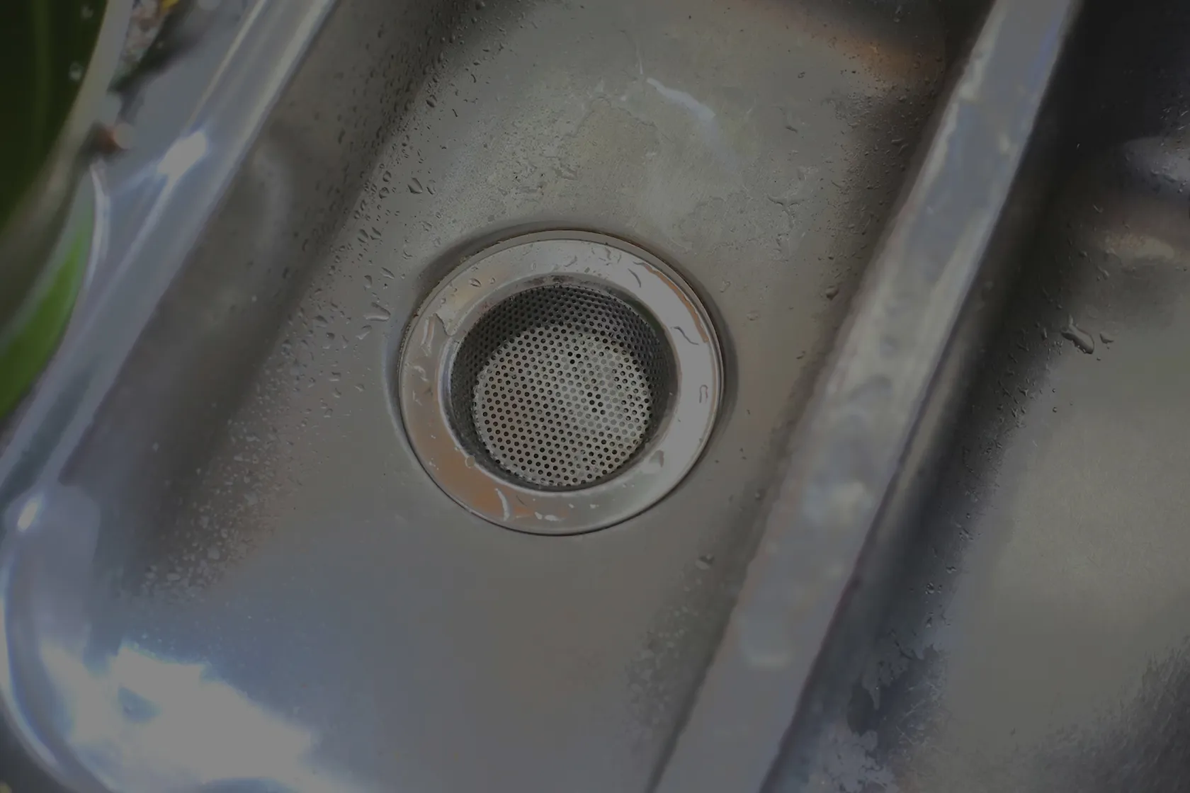 Close-up of a stainless steel kitchen sink drain with a perforated strainer and water droplets on the surface.