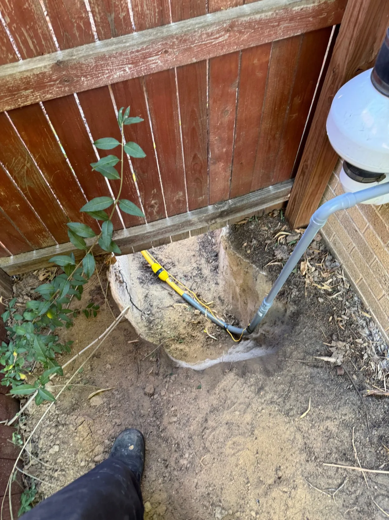 A partially dug hole next to a wooden fence and brick wall with exposed pipes and a yellow cable, with a person's black boot visible in the dirt.