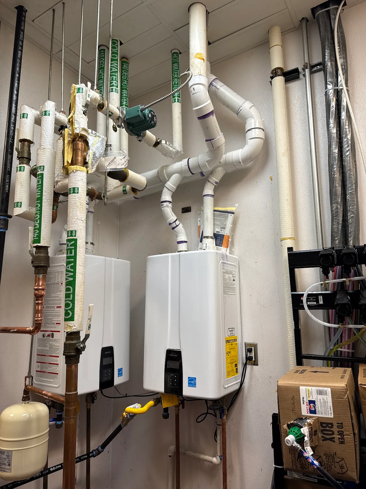Mechanical room with two white wall-mounted water heaters connected to multiple insulated pipes labeled 'cold water,' copper piping, a beige expansion tank, and a cardboard box on the right.