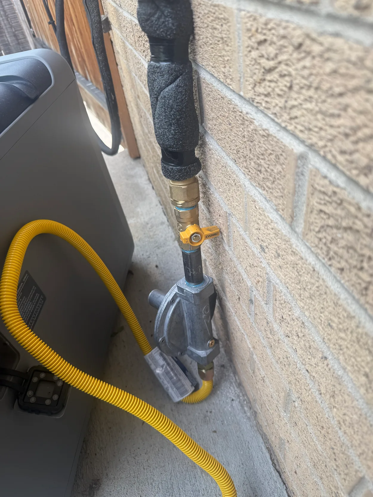 Outdoor gas line with a yellow corrugated flexible gas hose attached to a metal shut-off valve with a yellow handle mounted on a brick wall next to an air conditioning unit.