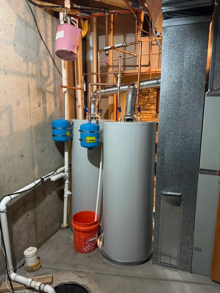 A basement corner with two tall water heaters connected to several copper pipes and two blue expansion tanks; an orange Home Depot bucket sits on the floor beneath one of the heaters, and white plumbing pipes run along the concrete wall and floor.