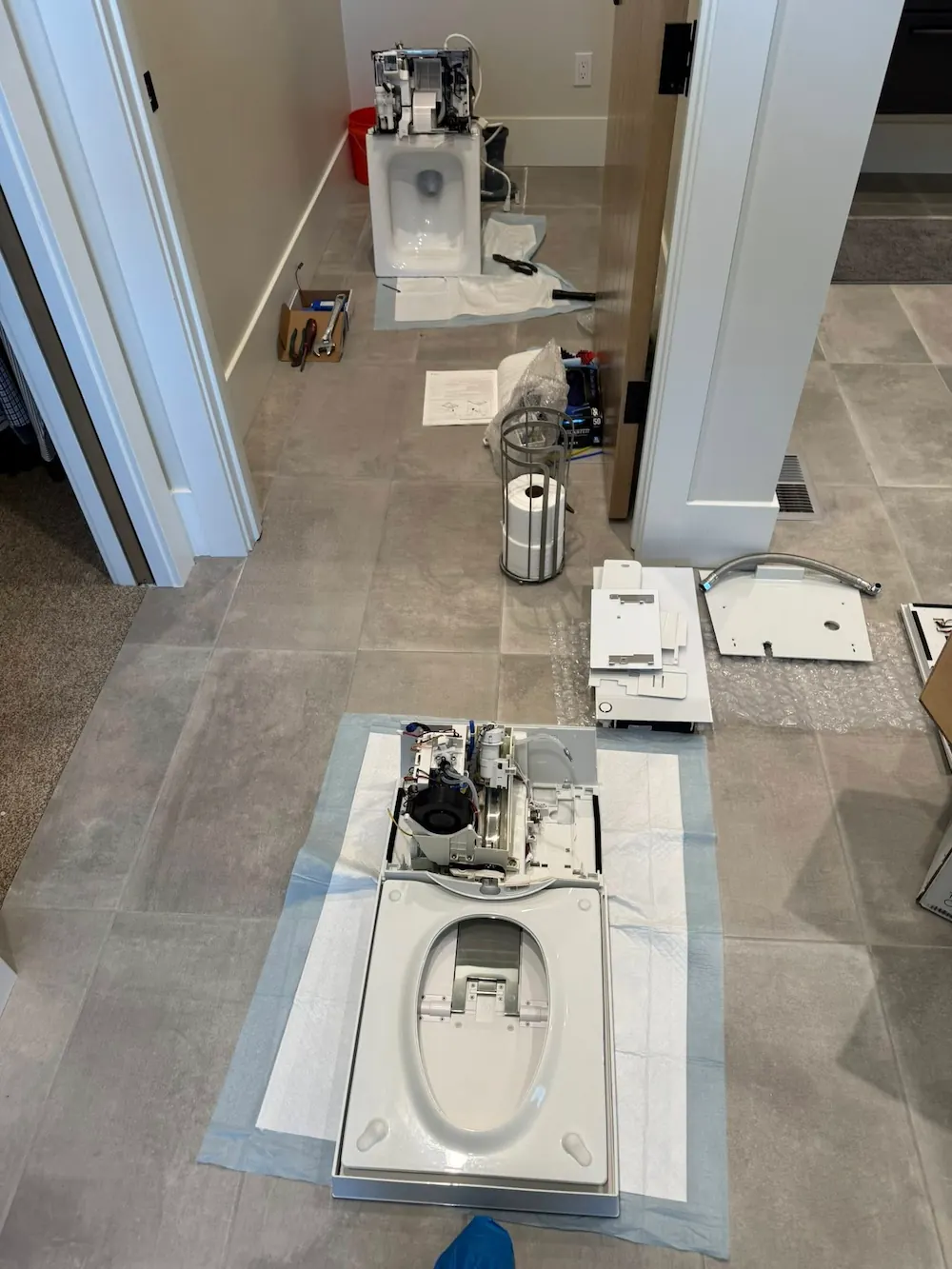 Unassembled smart toilet parts laid out on a tiled floor in a hallway, including two toilet bowls with exposed internal mechanisms, a toilet paper holder with a roll inside, various detached components, a box with tools, and instruction papers.