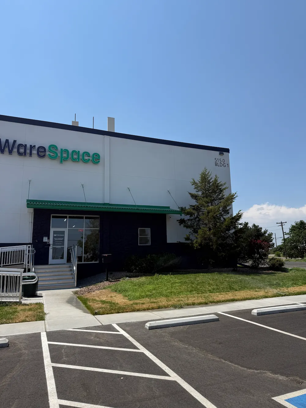 Exterior of a white commercial building with partial signage reading 'wareSpace' in black and green letters, a green awning above the entrance, wheelchair ramp and stairs, adjacent lawn, and parking spaces in front under a clear blue sky.