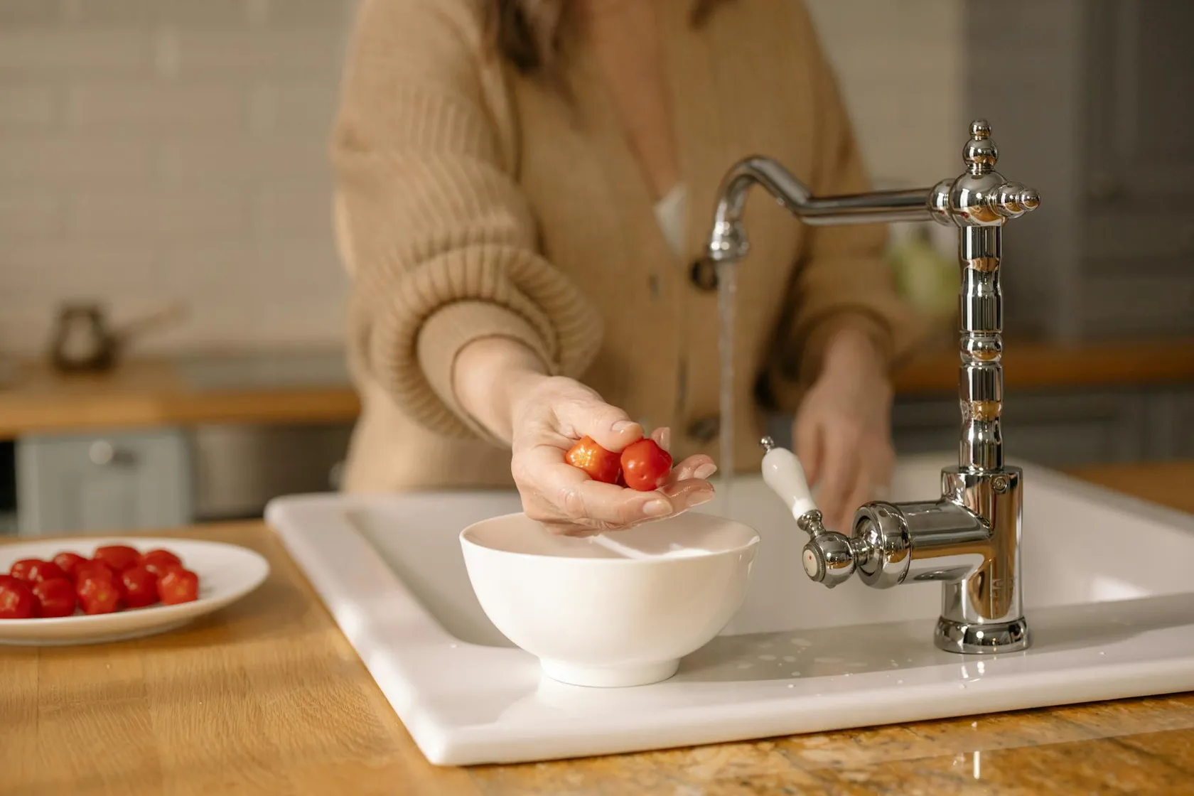 Person washing small red tomatoes under running kitchen sink faucet with a white bowl placed in the sink.