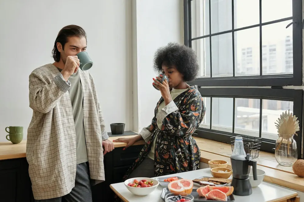 Man and woman standing in a kitchen by a window drinking from mugs, with a table set with bowls of fruit, sliced grapefruit, and a coffee maker.