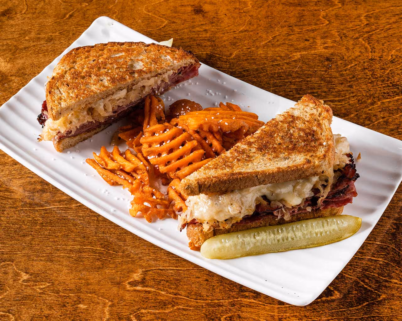 Reuben sandwich cut diagonally on a white rectangular plate with a side of waffle fries and a pickle spear, on a wooden table.