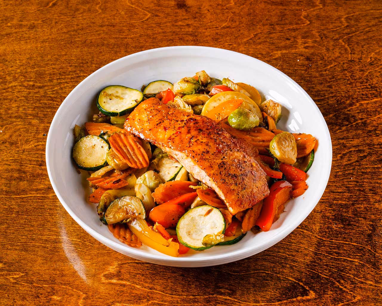 Grilled salmon fillet seasoned with spices served on a white plate with sautéed mixed vegetables including zucchini, carrots, Brussels sprouts, and bell peppers on a wooden table.