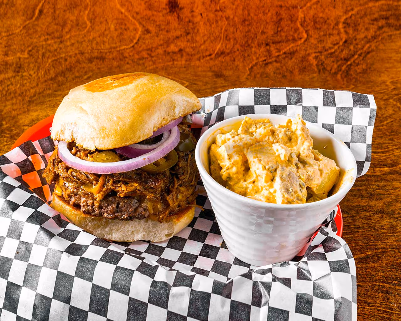 Pulled pork sandwich with slices of red onion and jalapeños on a toasted bun served with a side of creamy potato salad in a white cup on black and white checkered paper.