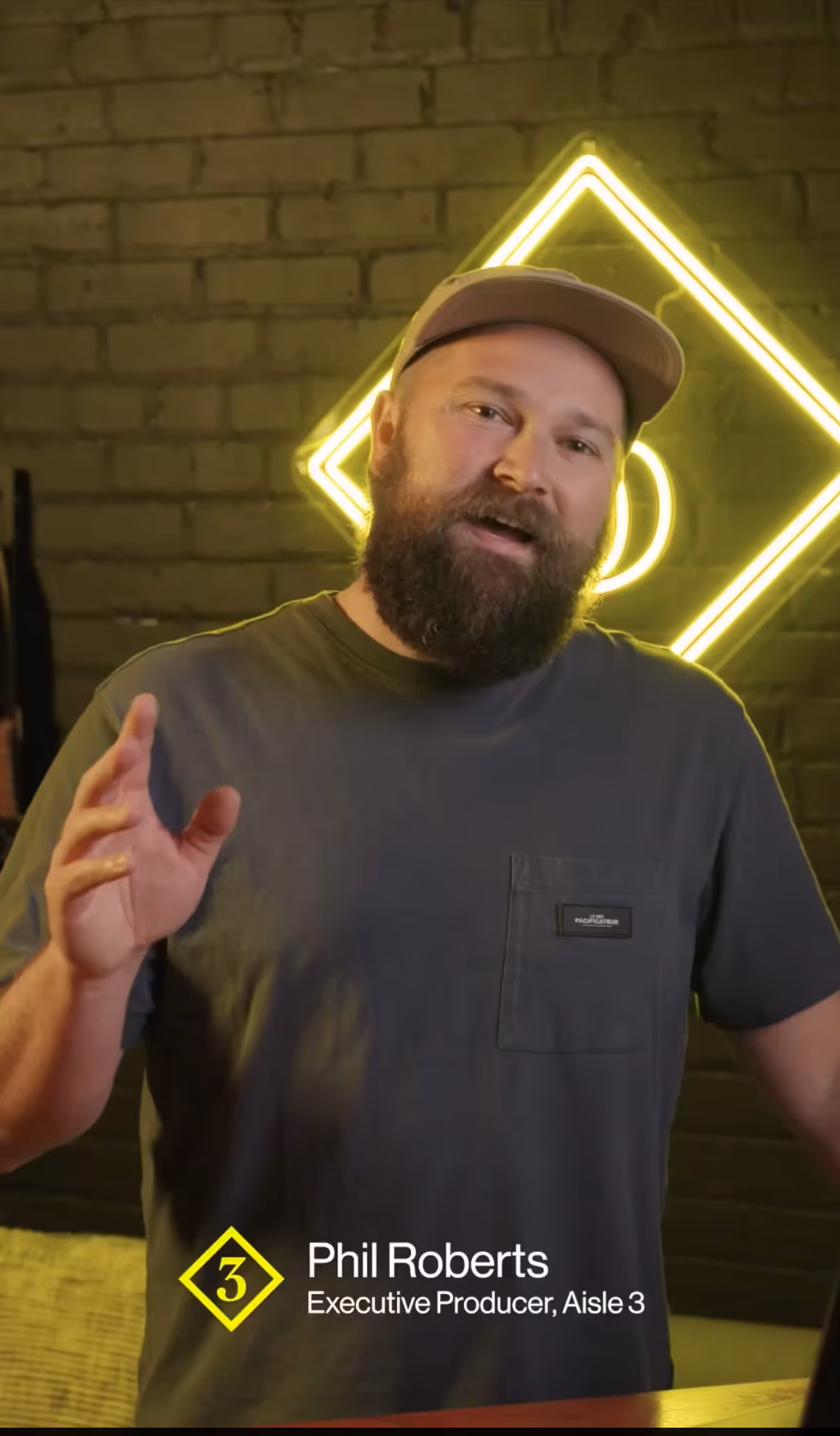 Bearded man wearing a cap and dark T-shirt speaking in front of a brick wall with yellow neon light, caption reads Phil Roberts, Executive Producer, Aisle 3.