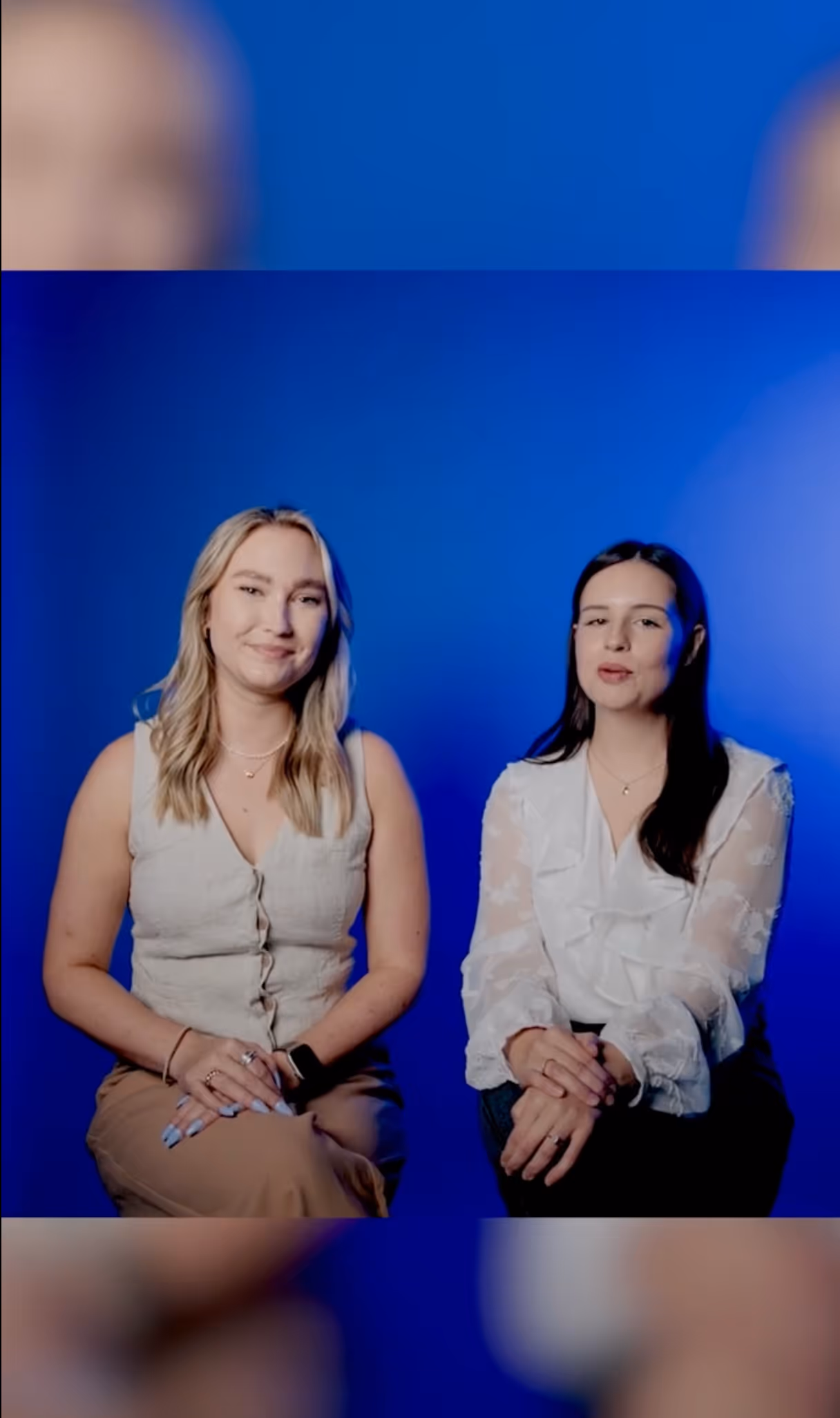 Two women sitting side by side against a blue background, one wearing a sleeveless beige top and the other in a white sheer blouse.