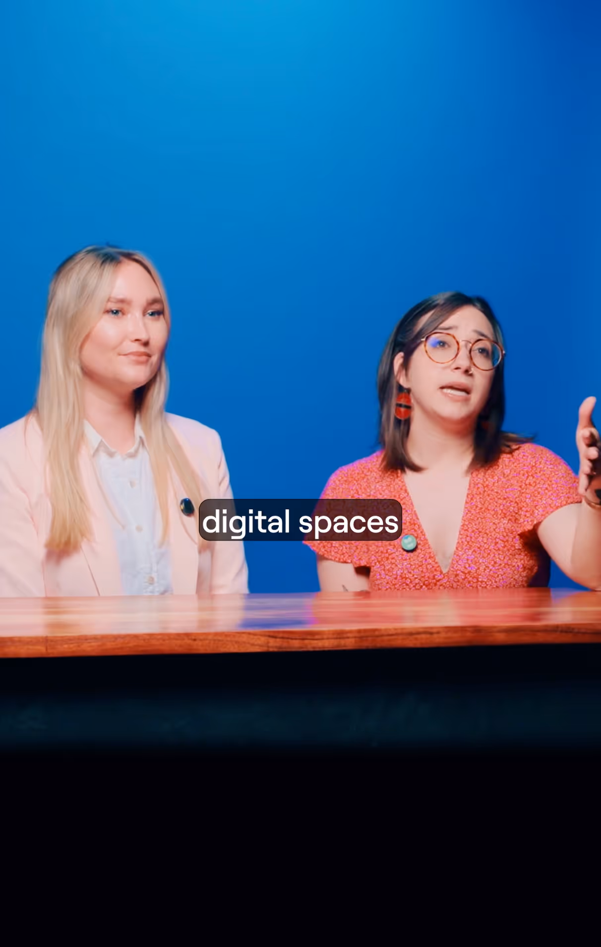 Two women sitting at a wooden table against a blue background, one speaking and gesturing with her hand, both wearing pins.