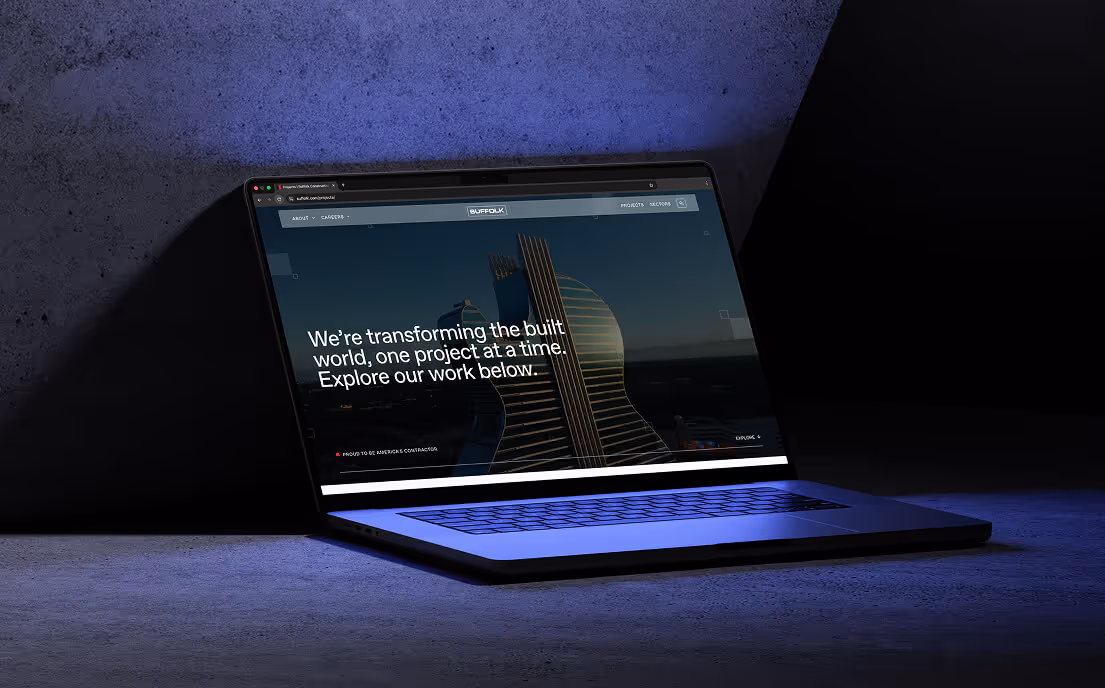 A laptop on a textured surface displaying a website with the text 'We're transforming the built world, one project at a time. Explore our work below.'